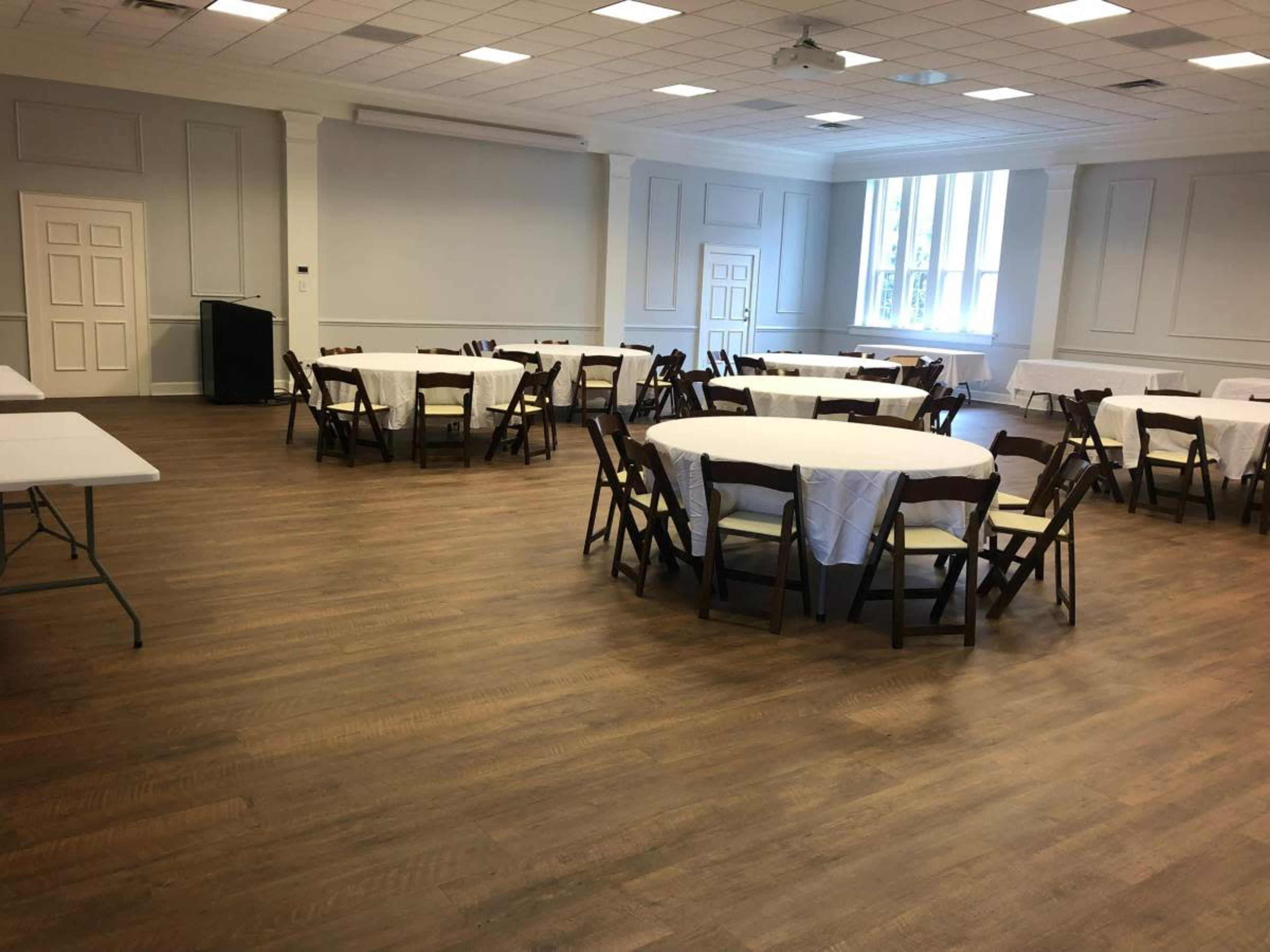 A large, empty event room with round tables covered in white tablecloths and wooden chairs arranged around them, along with a few folding tables along the side.