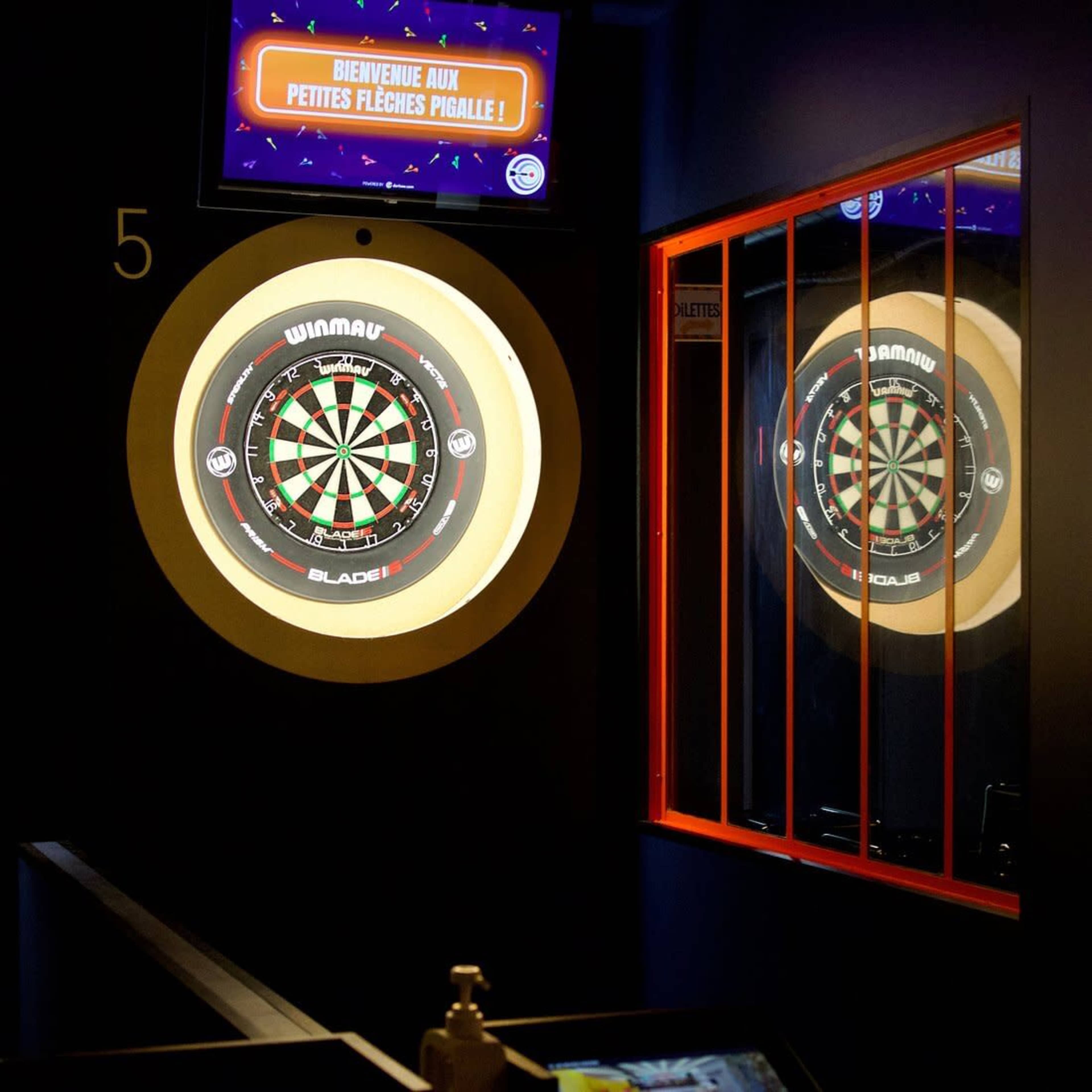 The image shows a dartboard illuminated in a dimly lit room, with a reflection visible in a nearby mirror.