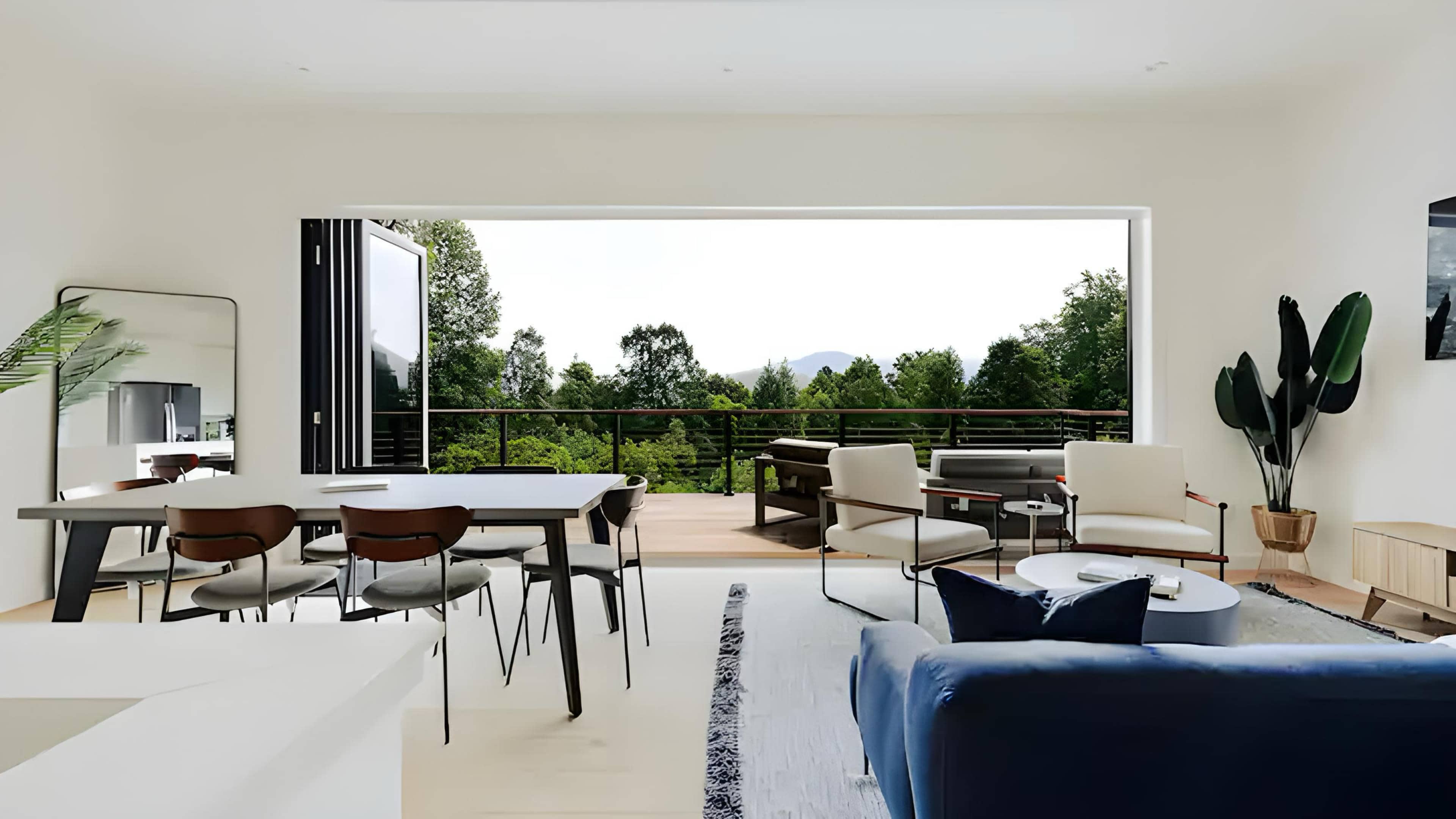 The living area features a large opening to a deck with views of a lush outdoor landscape, furnished with modern seating and a dining table.