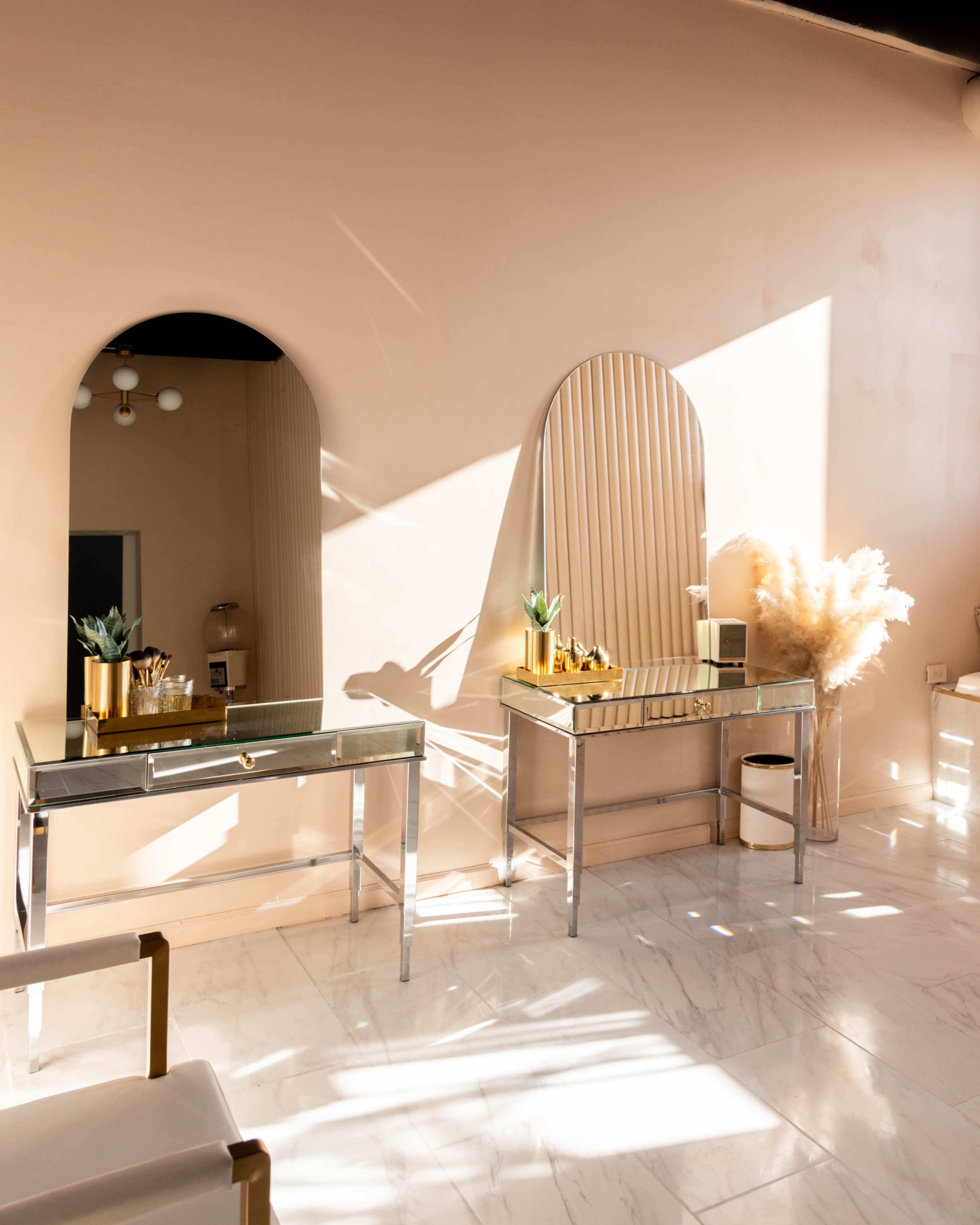 A bright interior featuring two sleek metallic tables with mirrors and decorative plants against a softly lit wall.