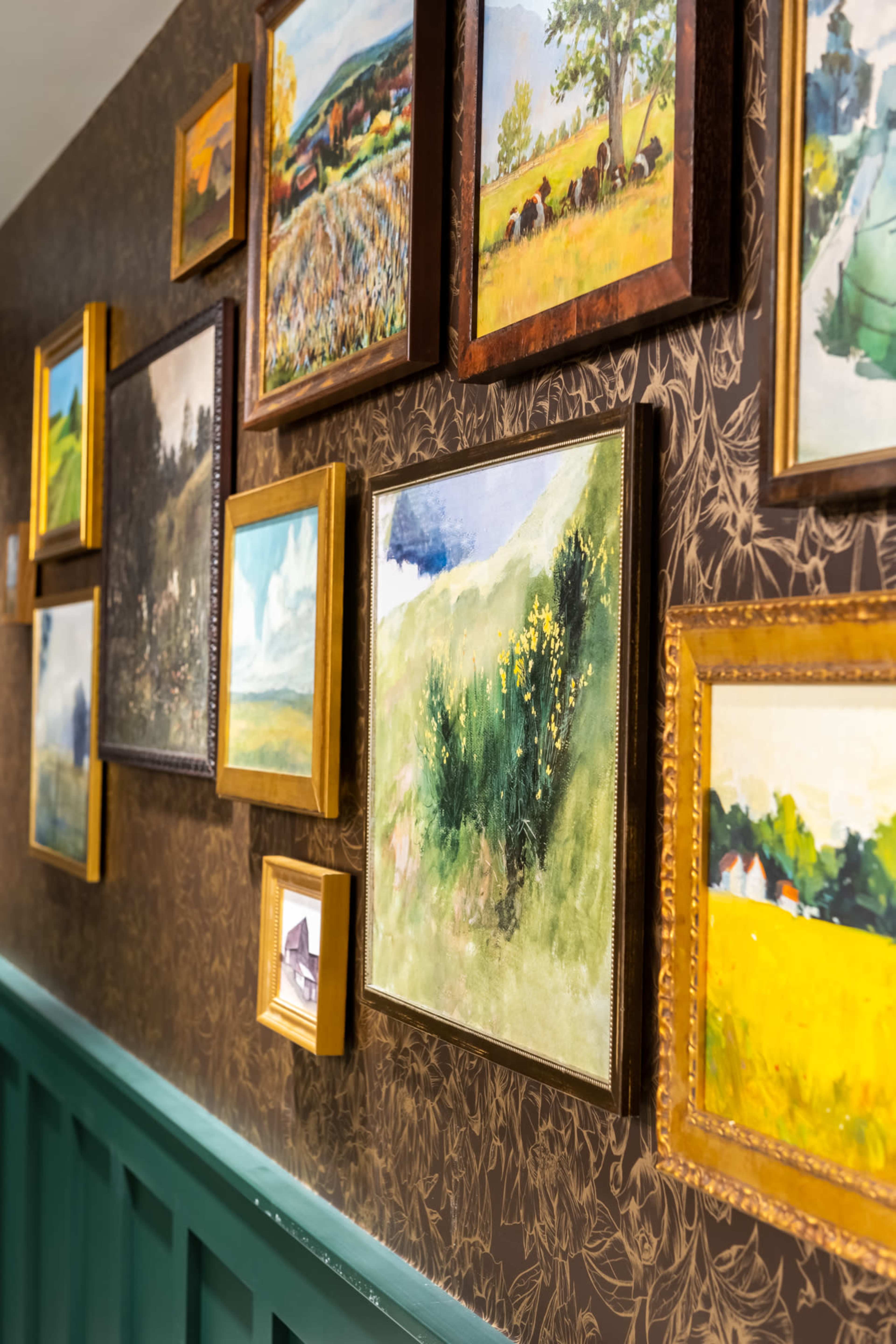 A wall adorned with various framed paintings displayed in a hallway, featuring landscapes and floral scenes.