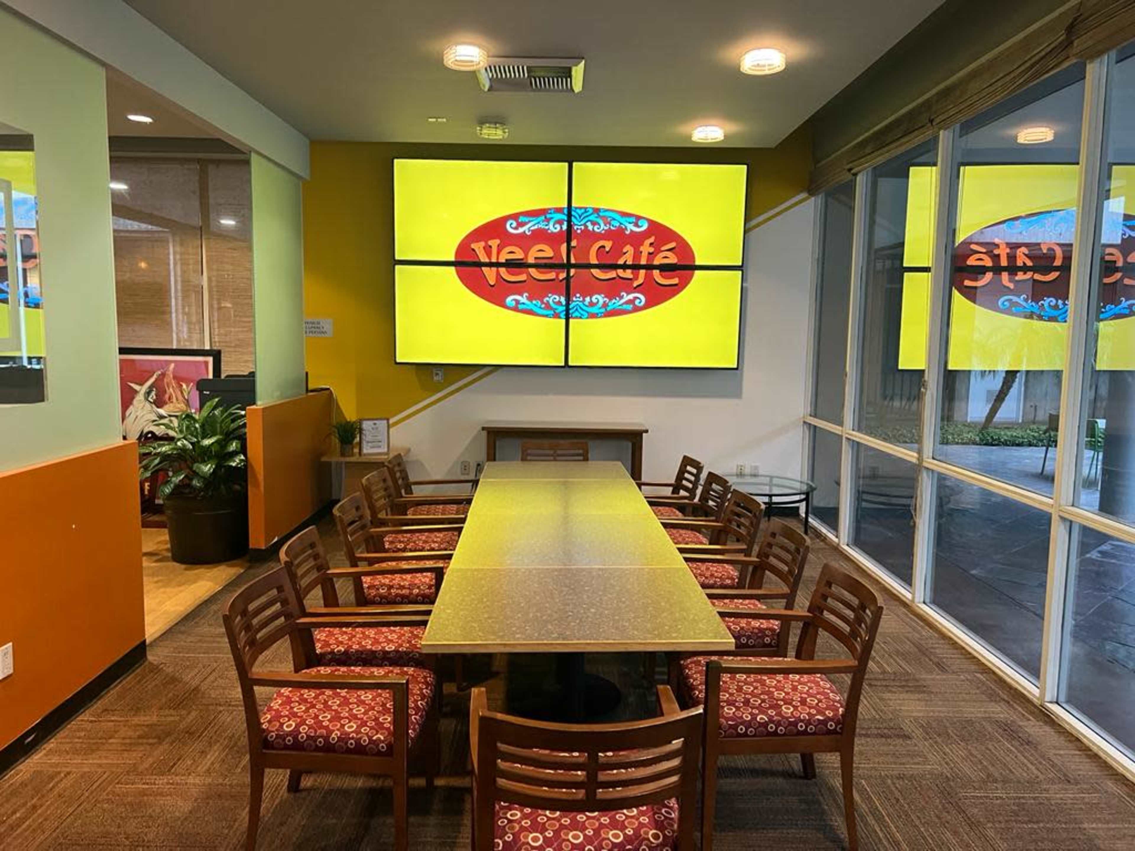A modern cafe meeting room featuring a long table surrounded by wooden chairs and large screens displaying the cafe's logo on the wall.