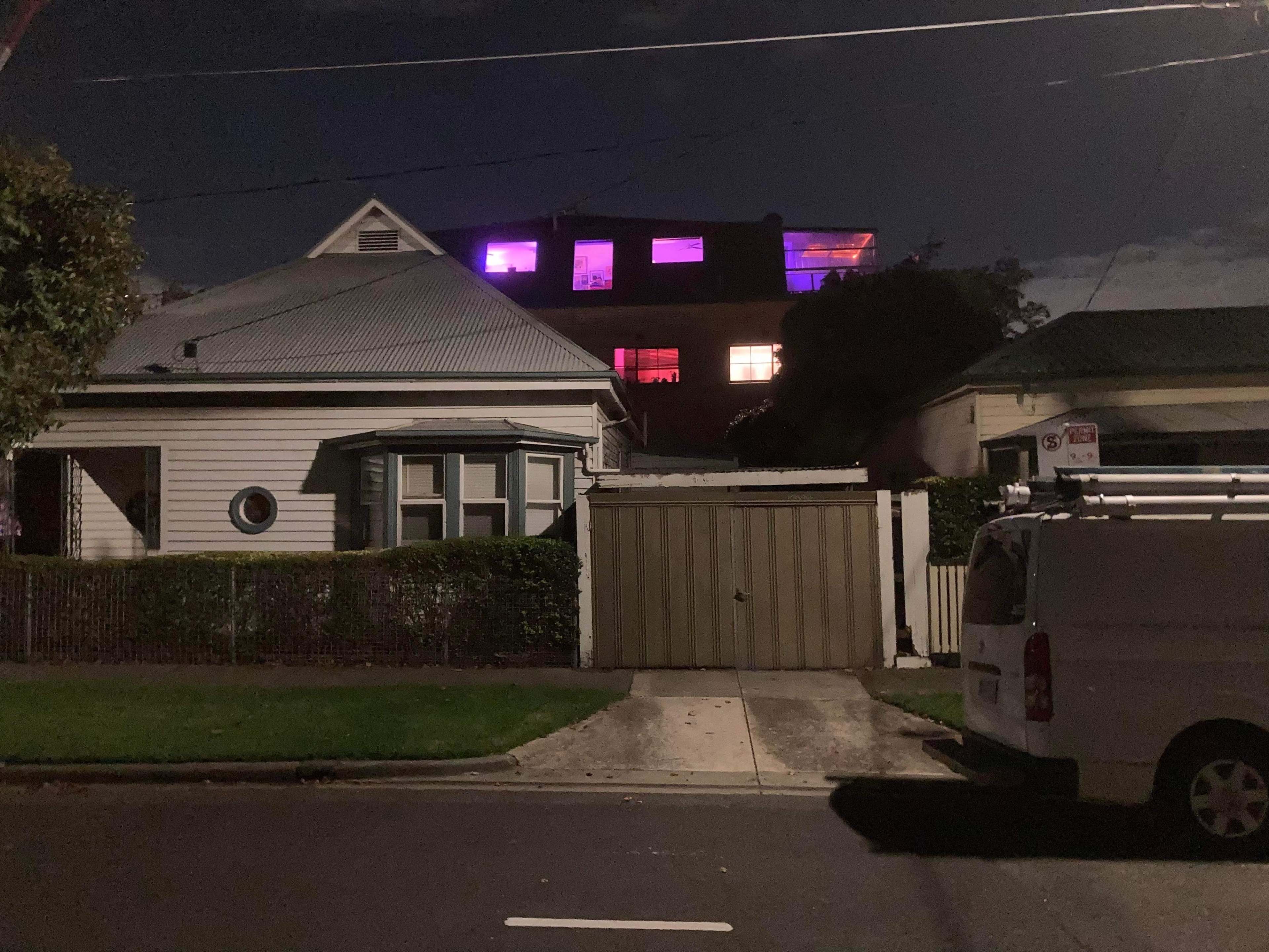A white house with a gray roof stands beside a garage, while a larger building behind it features illuminated windows in purple and red hues at night.