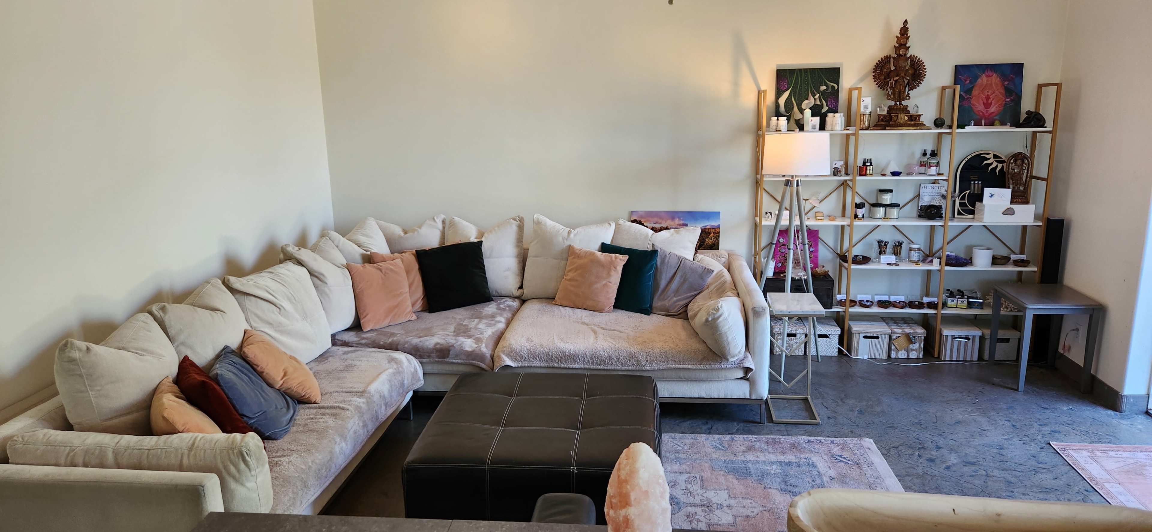 A spacious living area features a large sectional sofa with various cushions, a dark ottoman, and a shelf displaying decorative items and small products.