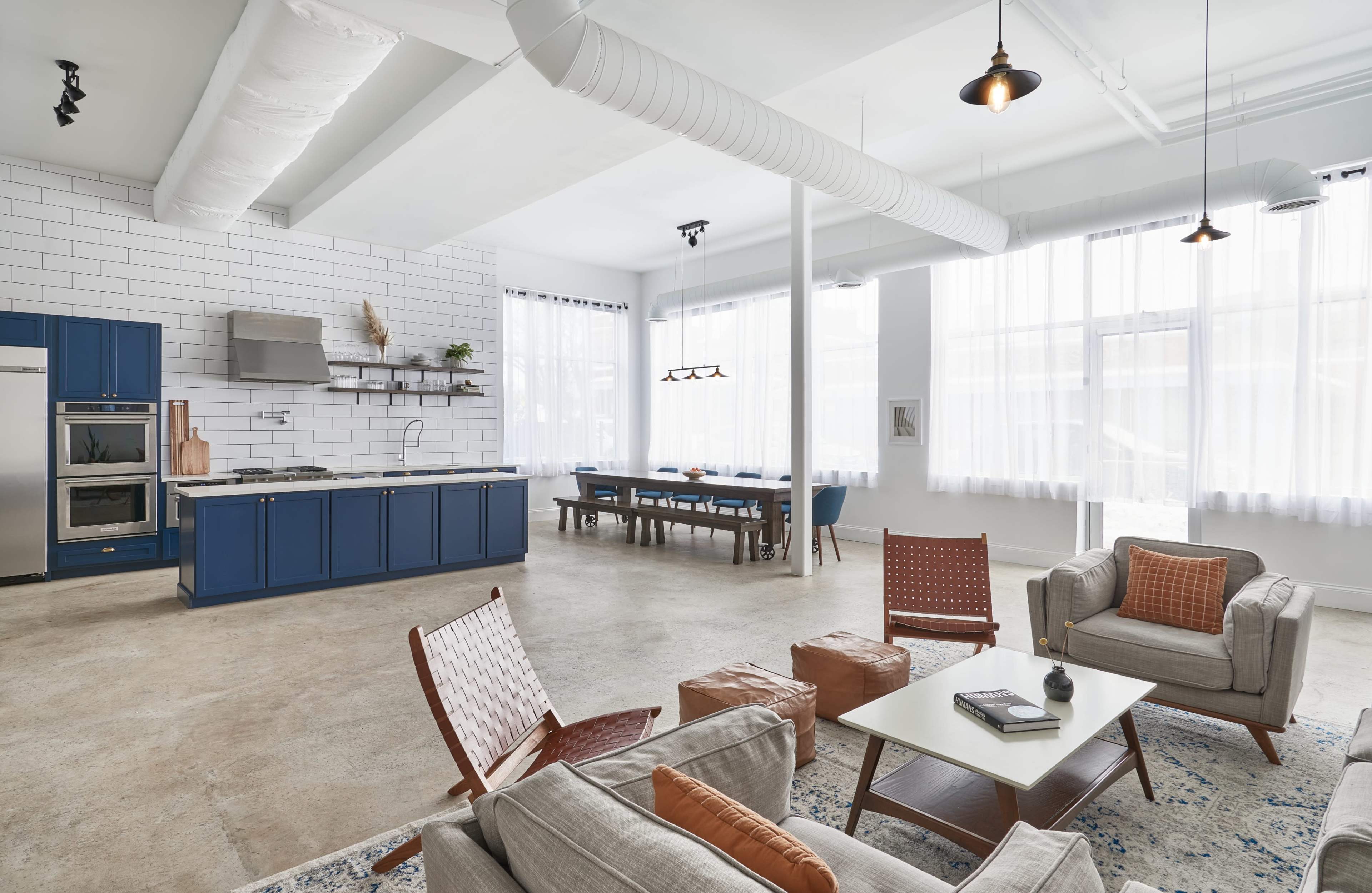 A spacious modern kitchen and living area features blue cabinetry, a large dining table, and ample natural light from tall windows.