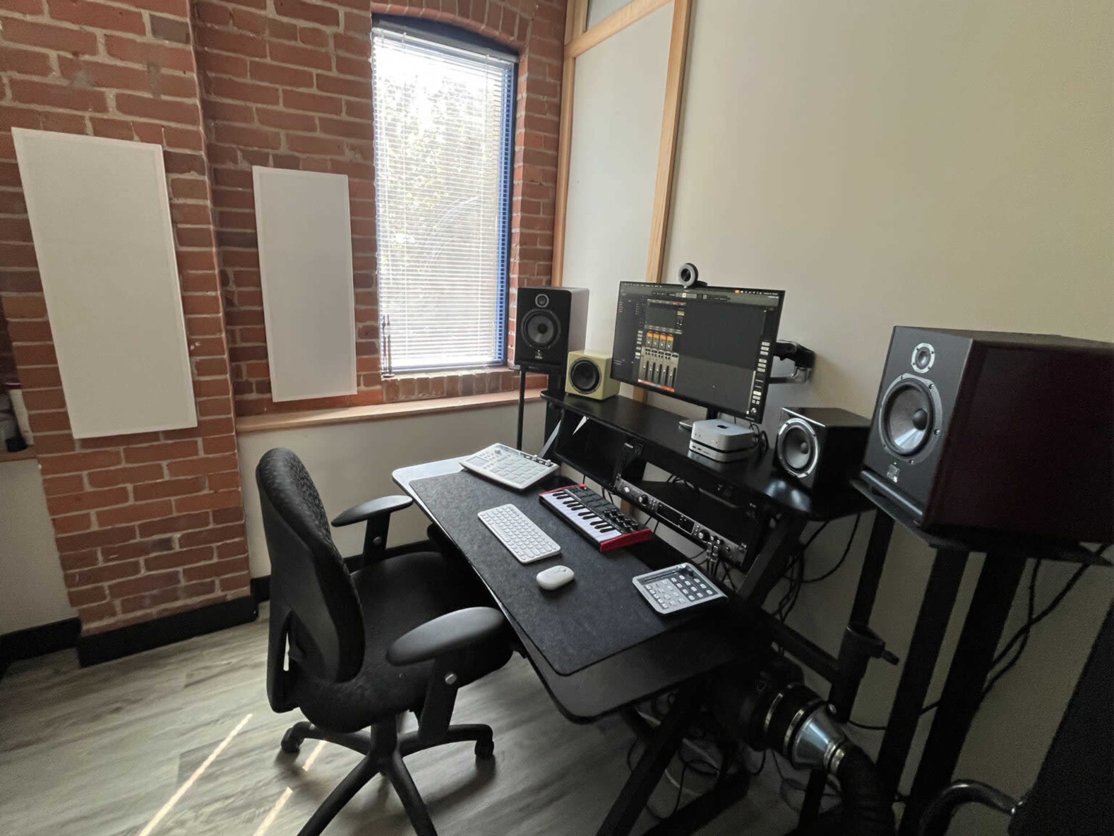 The image shows a music production studio with a desk, computer, and sound equipment placed against a brick wall.