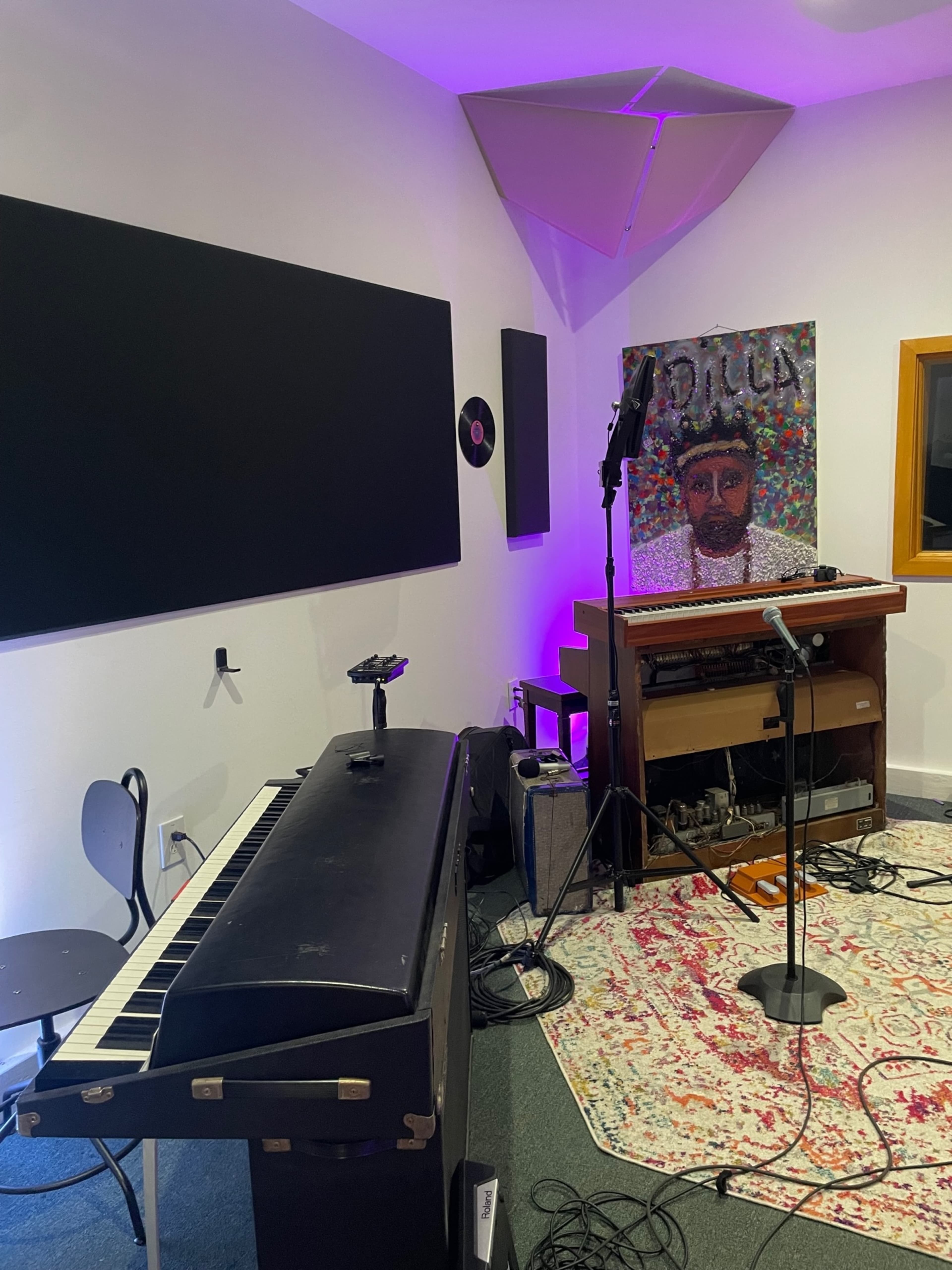 A music studio features a vintage keyboard, a microphone stand, and a colorful portrait of a man on the wall, accompanied by audio equipment and a patterned rug.