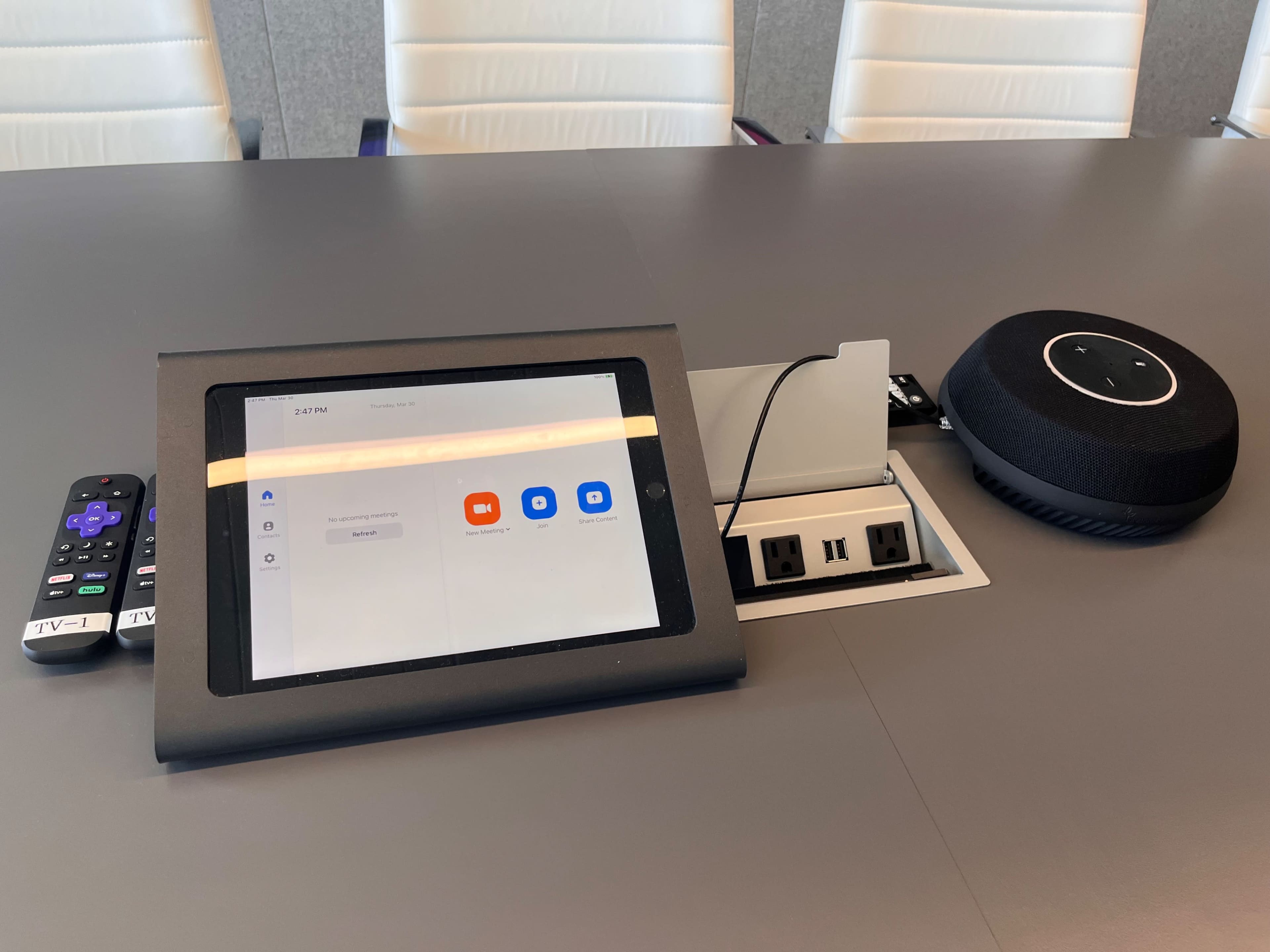 A tablet displays video conferencing options next to a remote control and a speaker in a modern conference room setup.