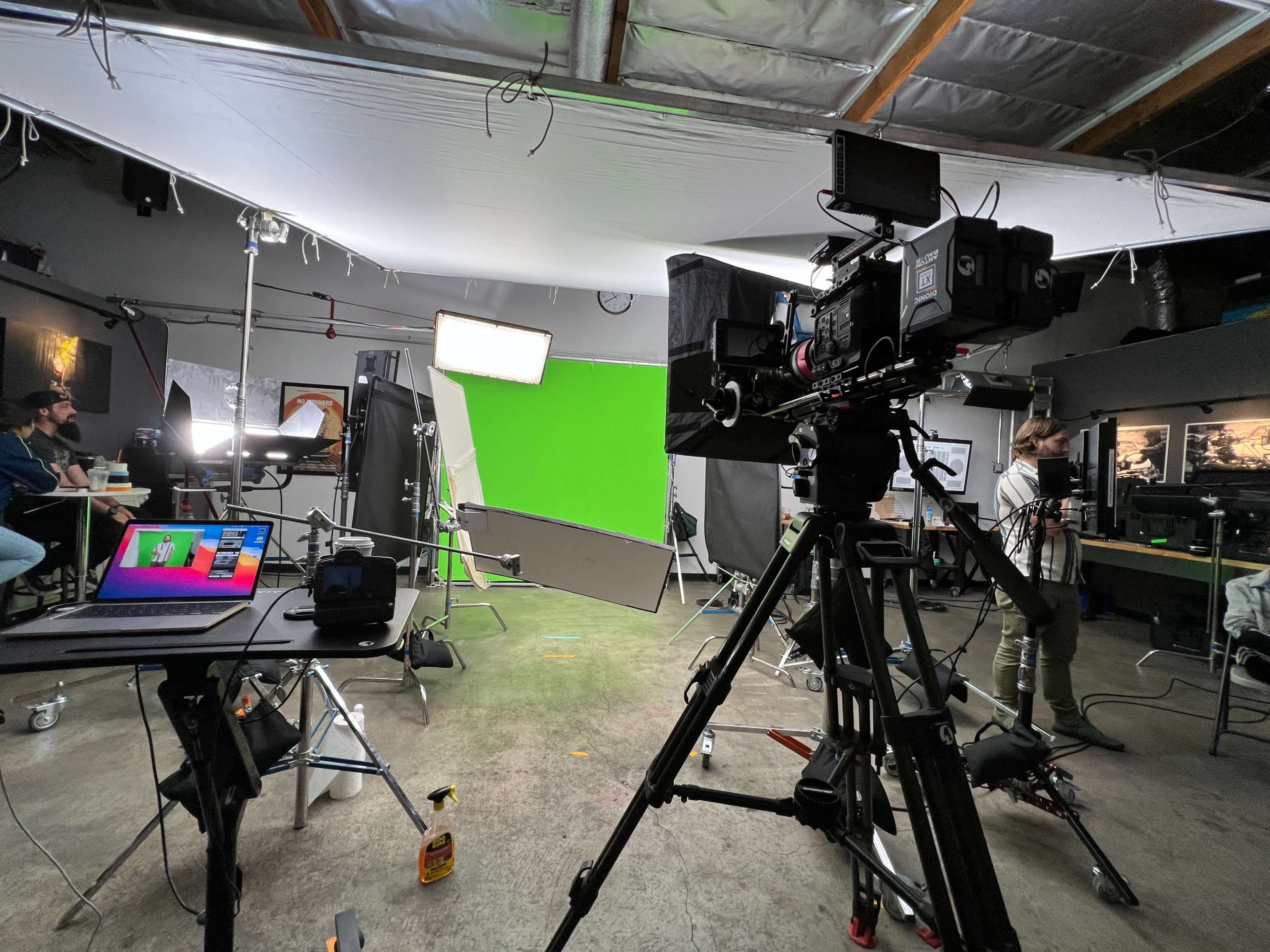 A film set is equipped with a green screen, lighting rigs, and cameras, while crew members prepare in the background.