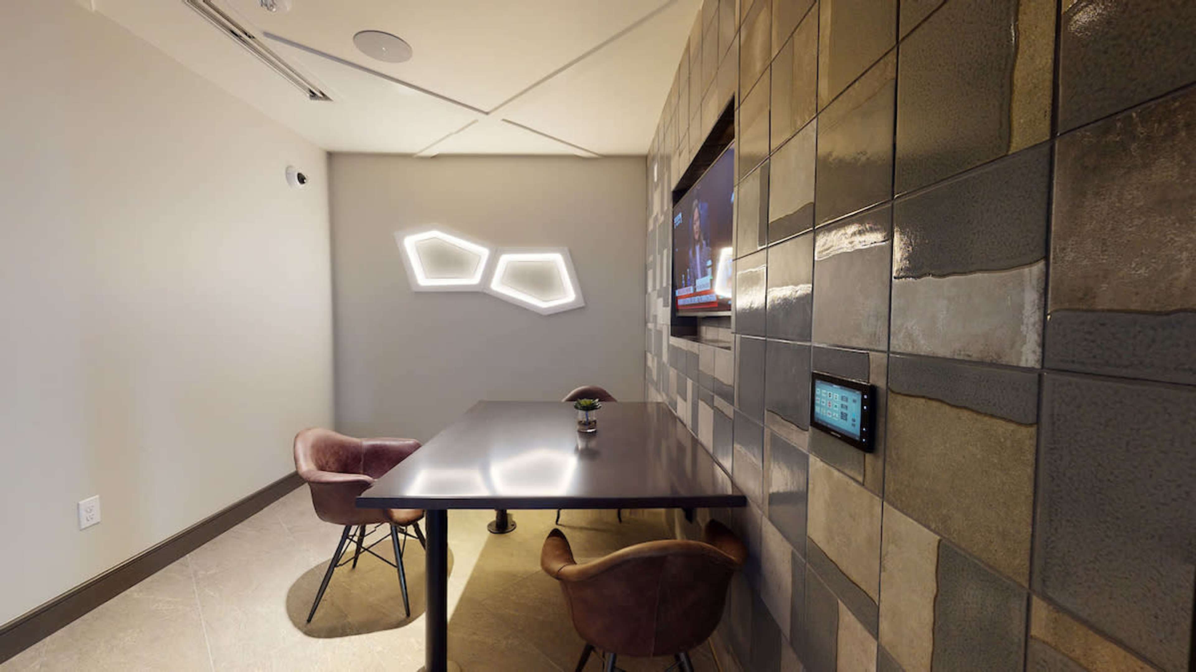 A small meeting room with a dark table, two chairs, a wall featuring geometric decorative lights, and a television mounted on the wall.