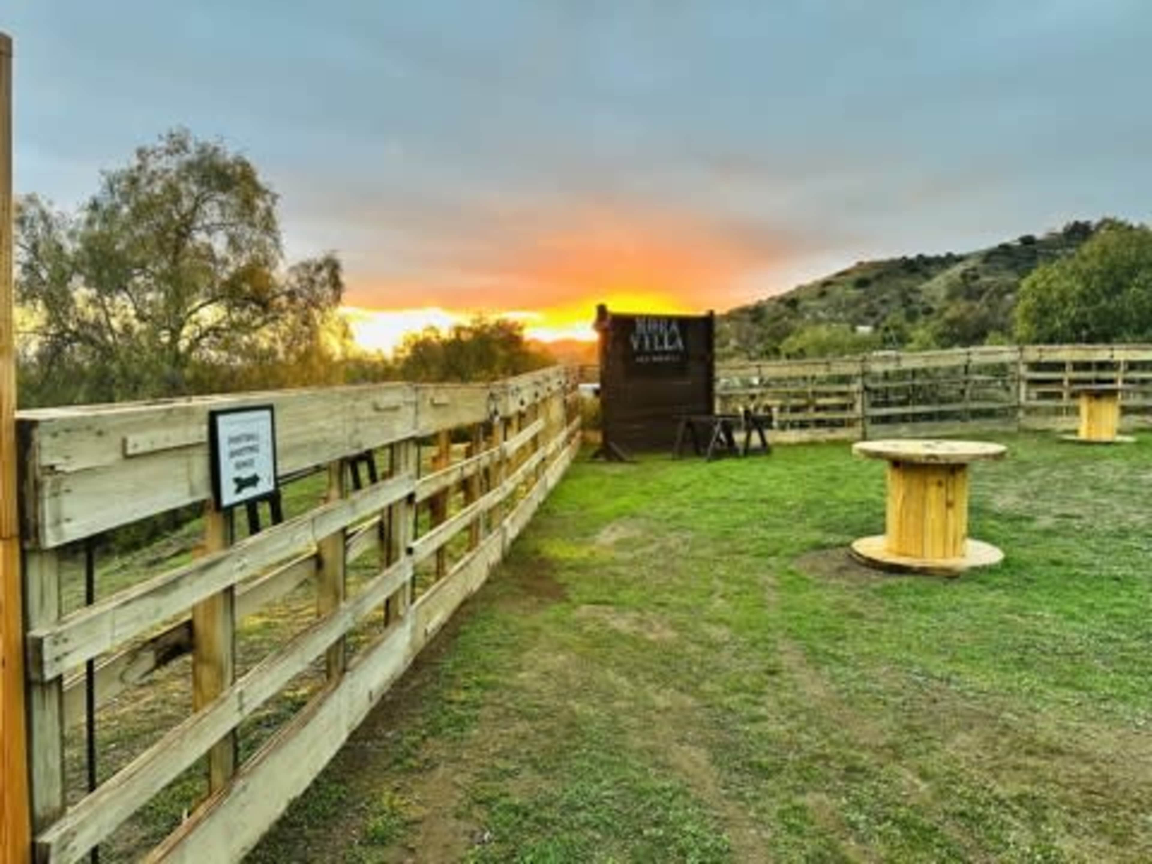 The image shows a wooden fence enclosing an outdoor area with grassy ground and a sunset visible in the background.