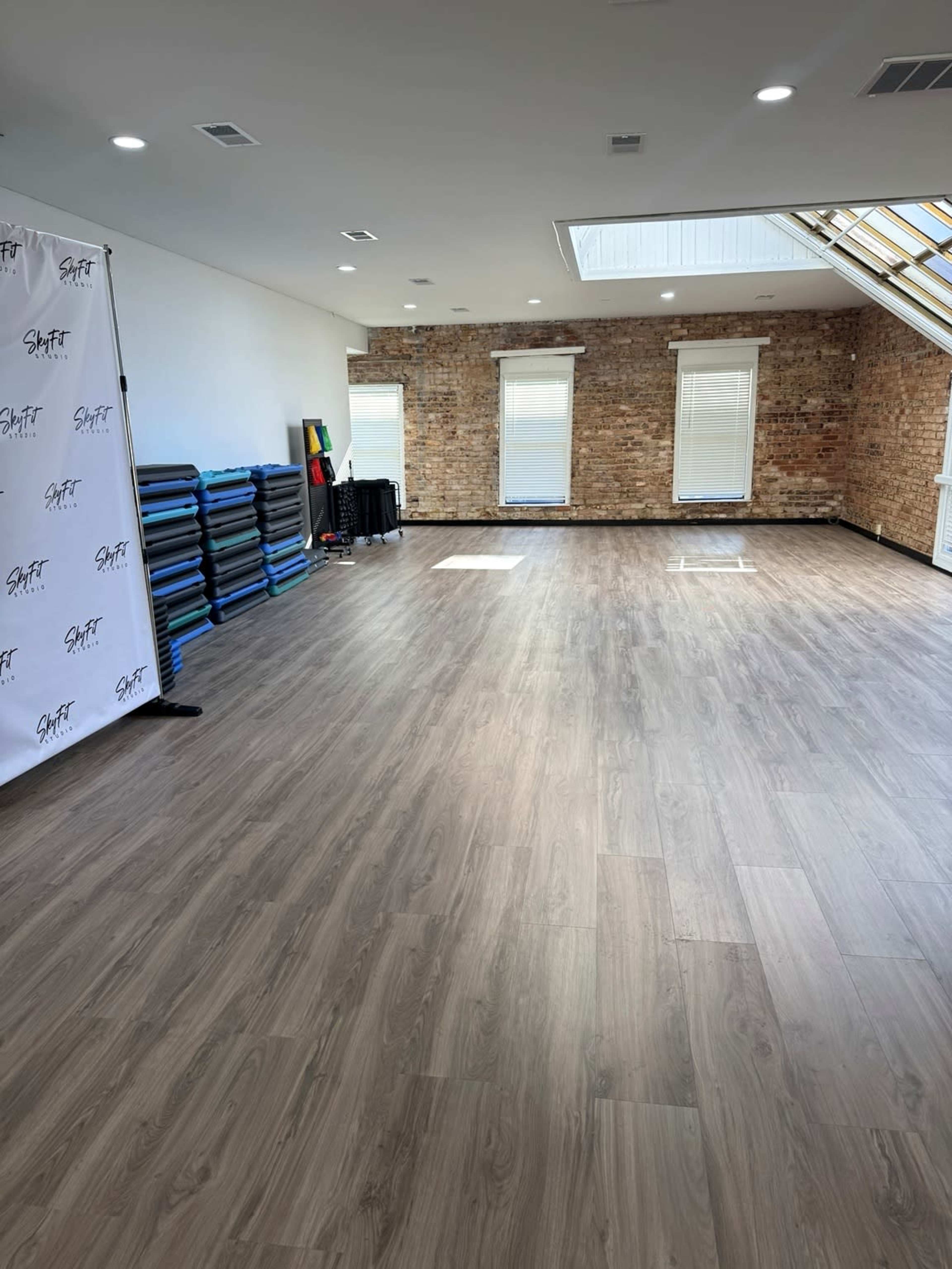 The image shows an empty fitness studio with wooden flooring, brick walls, and large windows.
