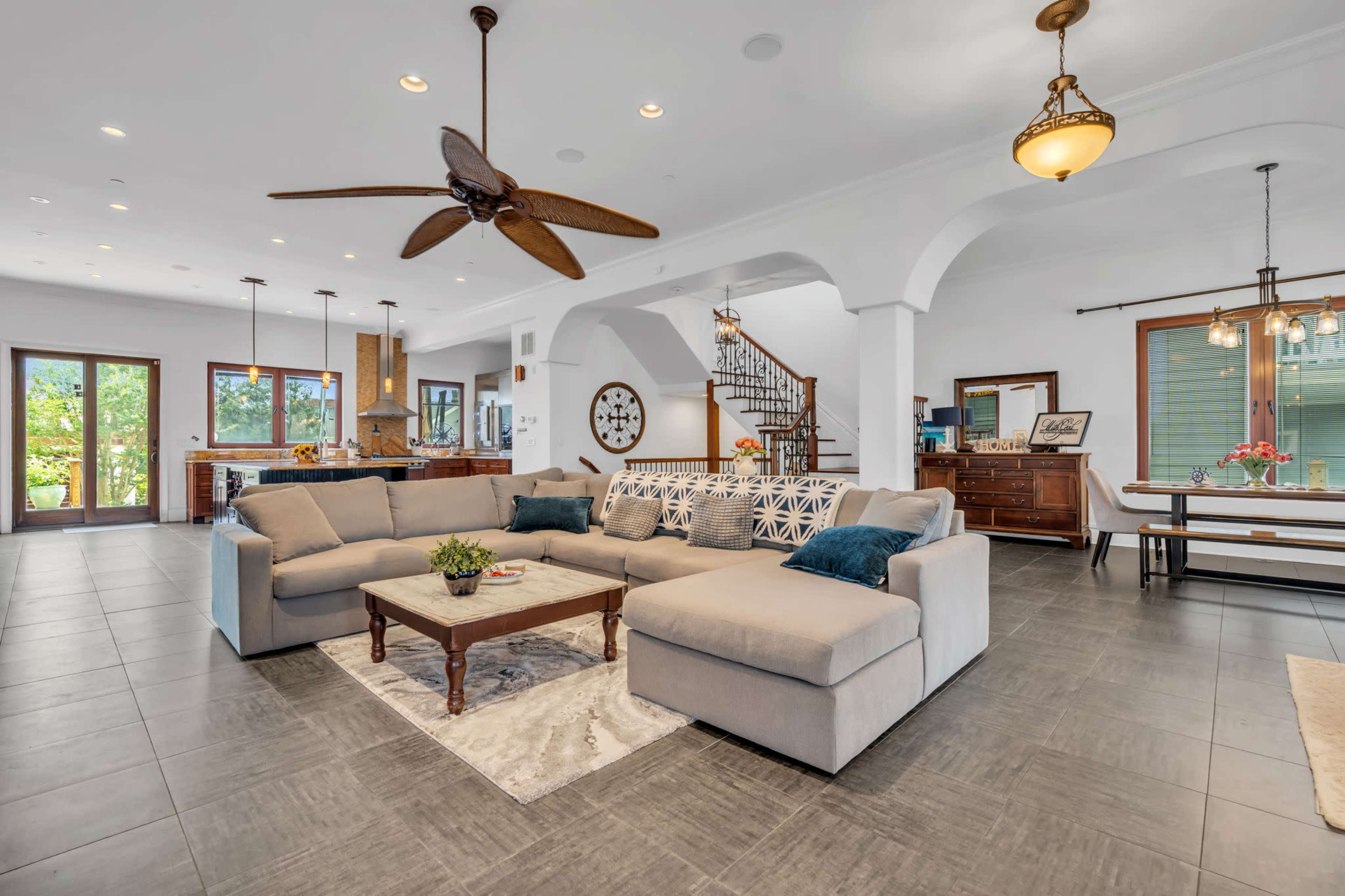 The image shows a spacious living room with a large sectional sofa, a central coffee table, and a staircase leading to another level, all set in a bright area with windows and modern decor.