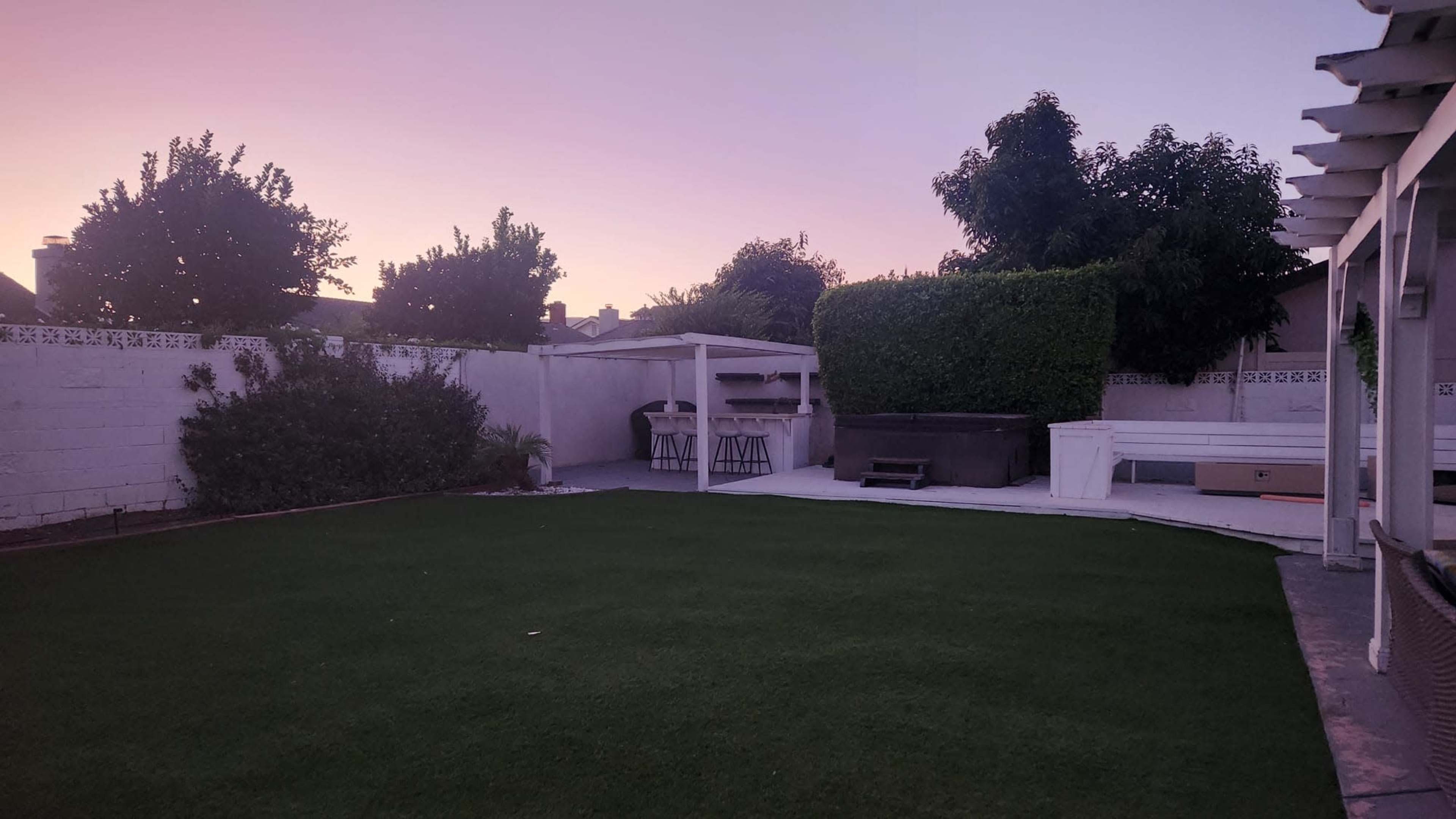 The image shows a backyard with artificial grass, a covered outdoor kitchen area, and trees against a pastel-colored sky at dusk.