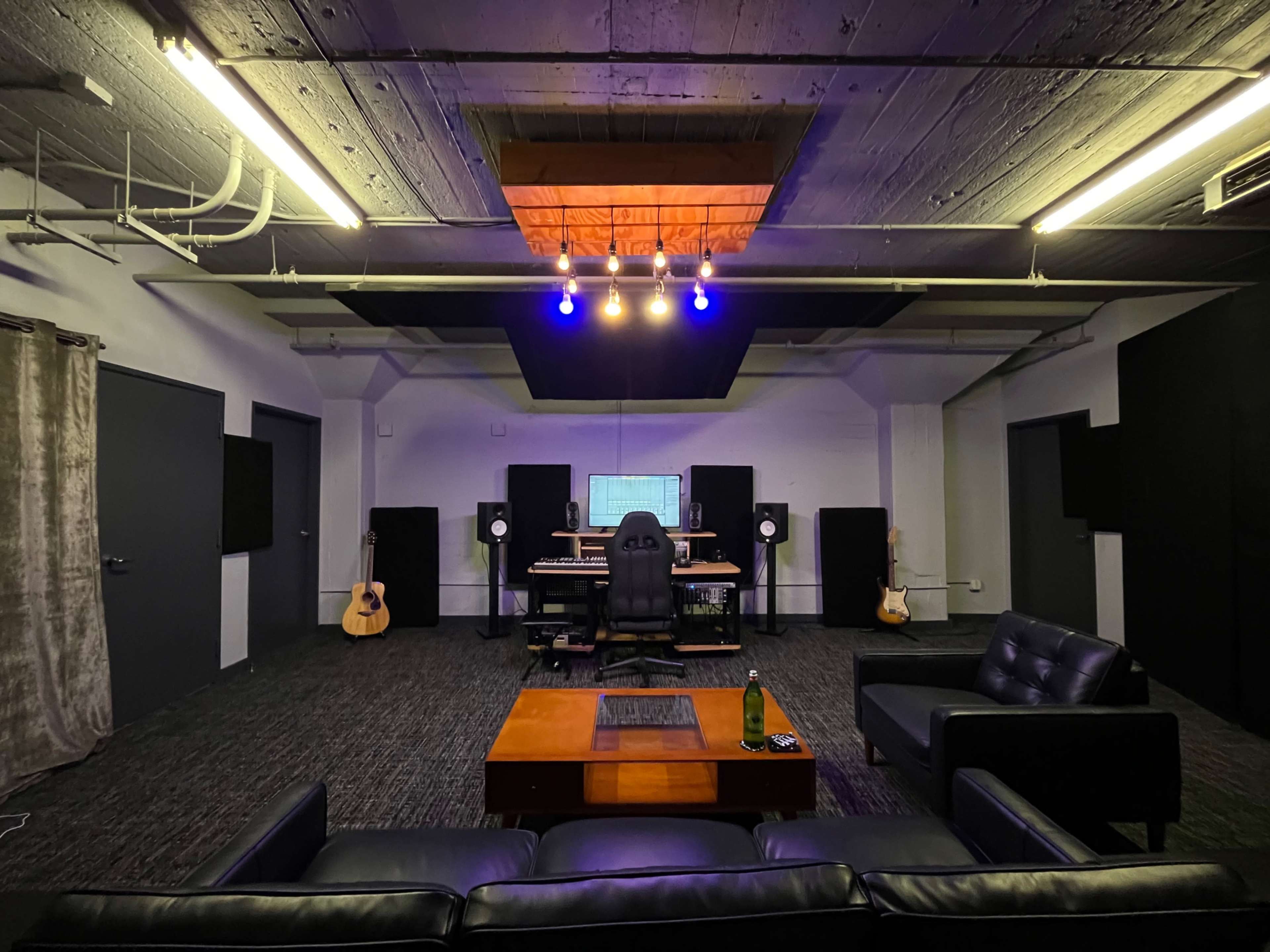 The image shows a music studio featuring a central control desk with audio equipment, surrounded by acoustic panels, two guitars, and a comfortable seating area.