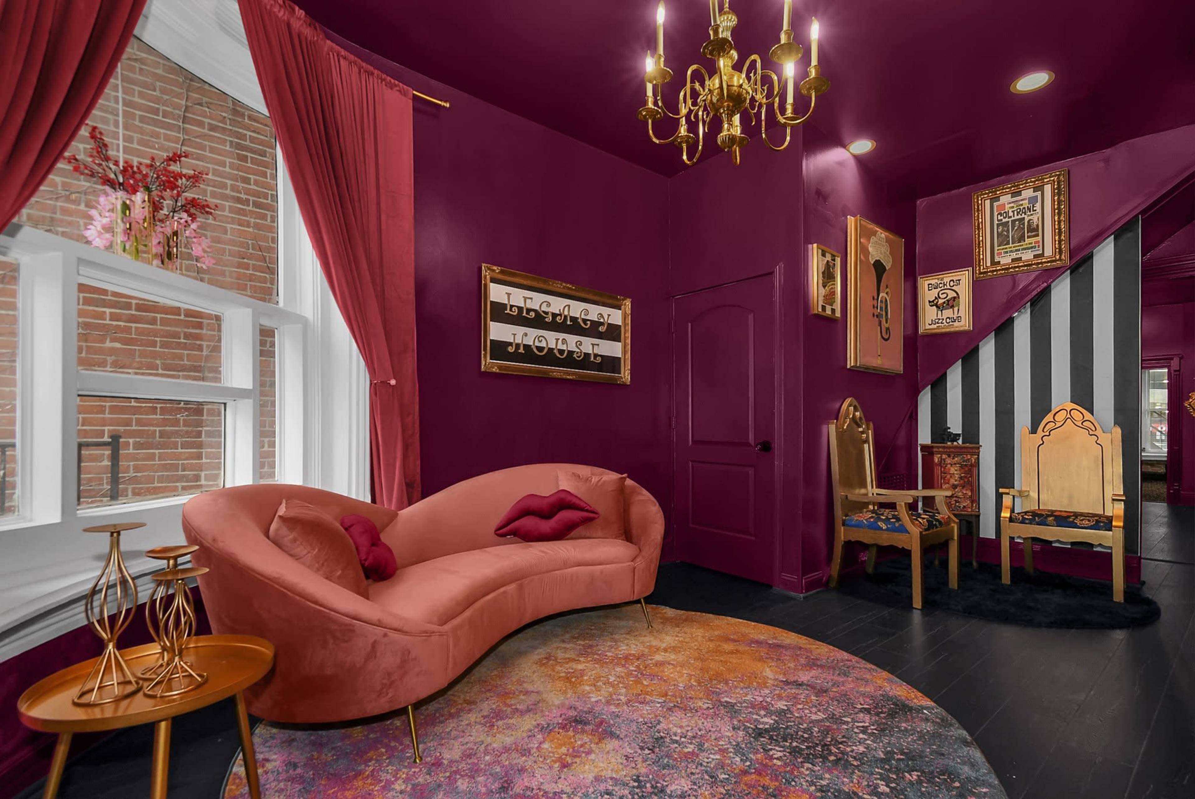 A richly colored room with a curved pink couch, a round rug, a chandelier, and decorative wall frames, featuring a staircase in the background.