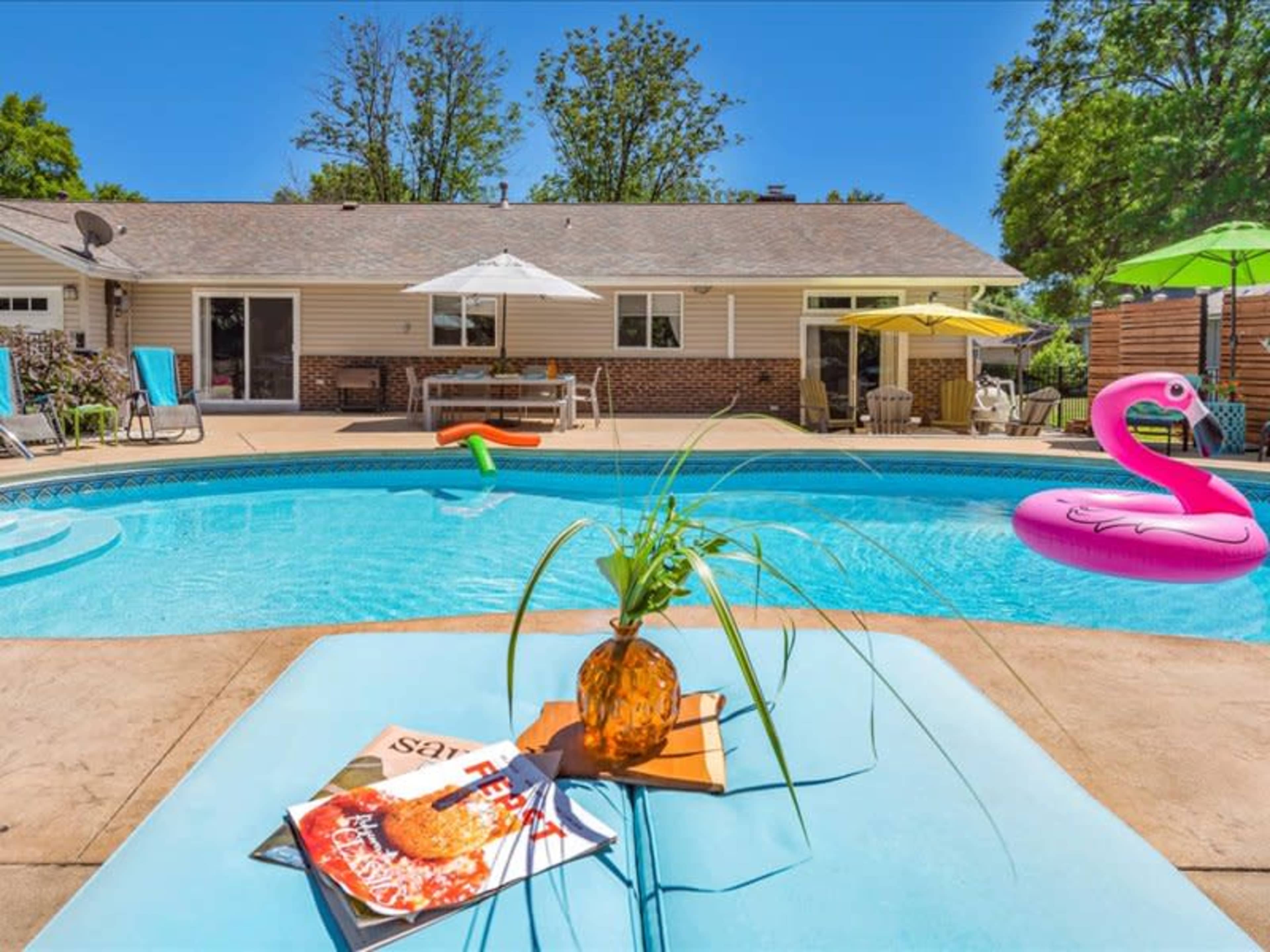 A backyard swimming pool with lounge chairs, a table with a magazine, and a pink inflatable flamingo floating on the water.