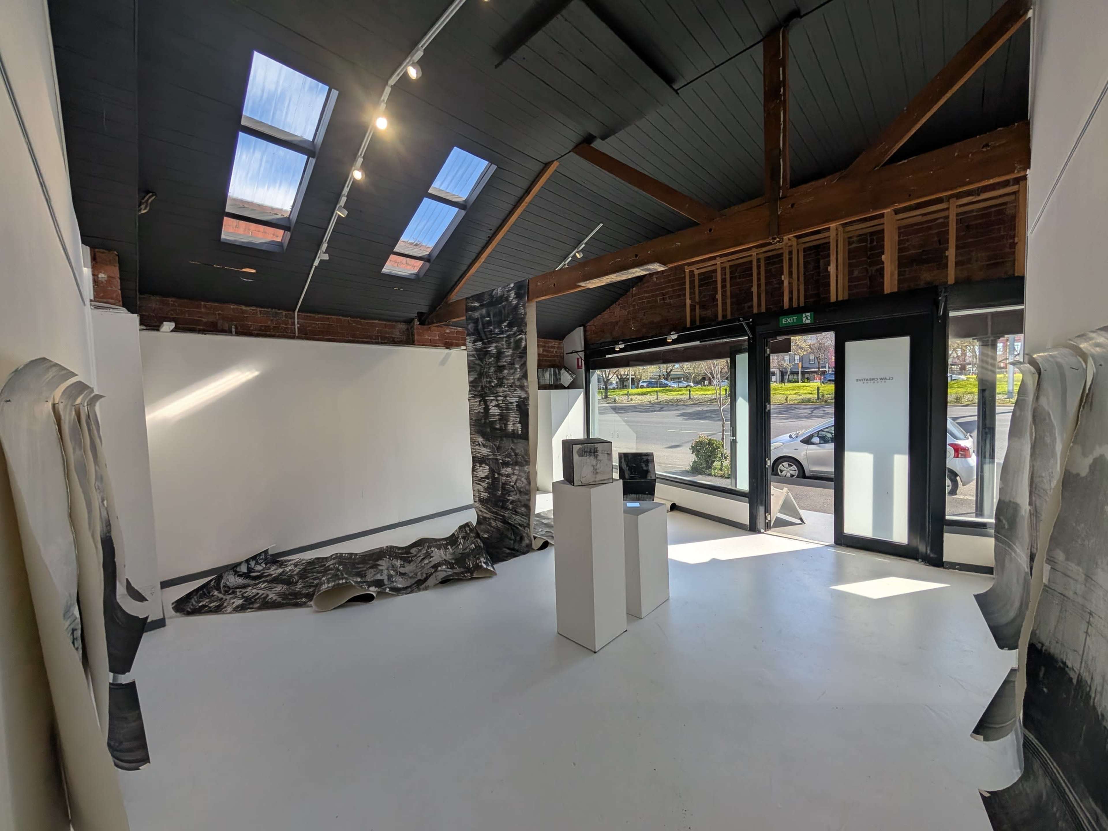 The image depicts a spacious, modern art gallery with white walls, wooden beams, and large windows allowing natural light to illuminate the interior.