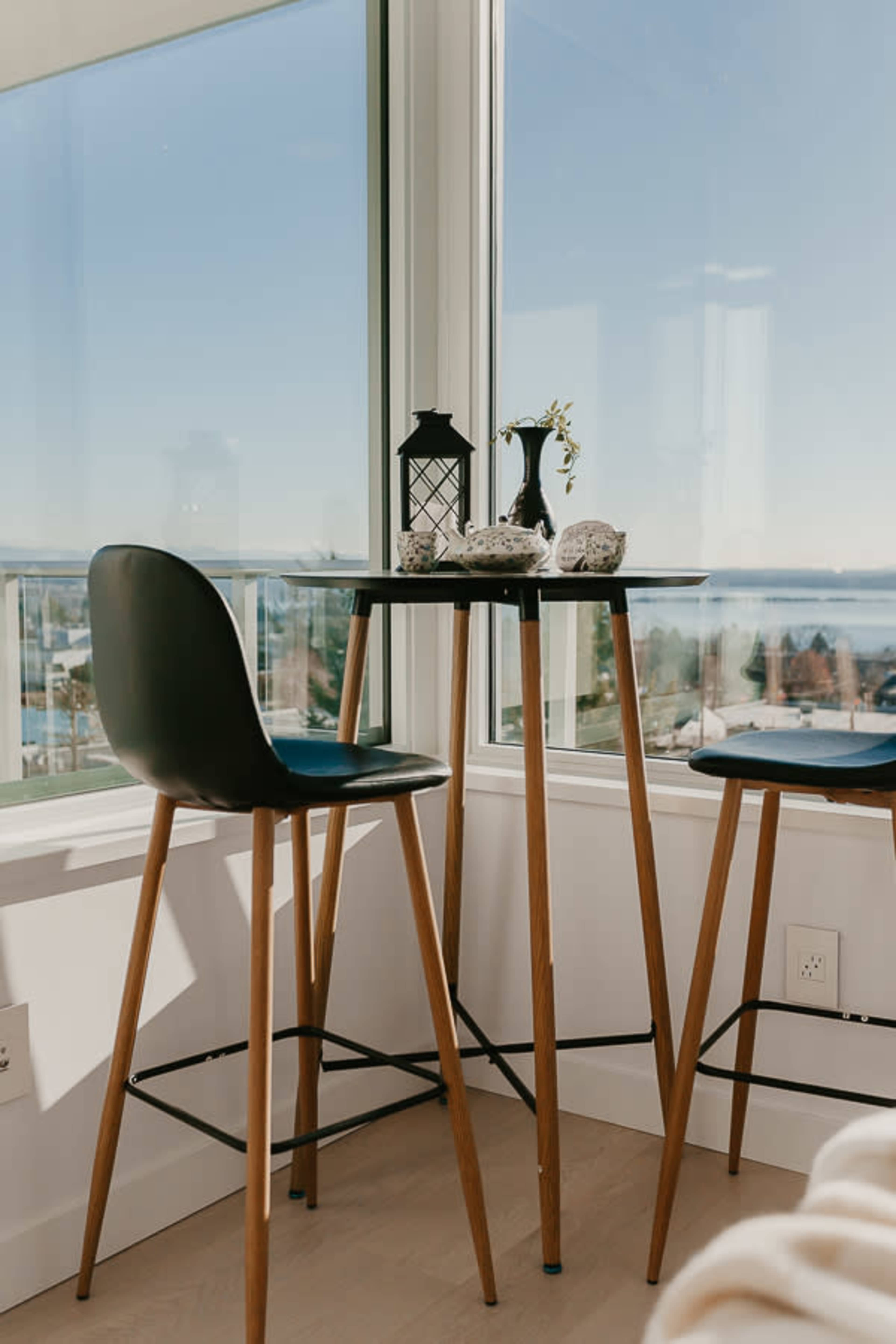 A small round table with two tall chairs is positioned by a large window, overlooking a scenic view of water and hills.