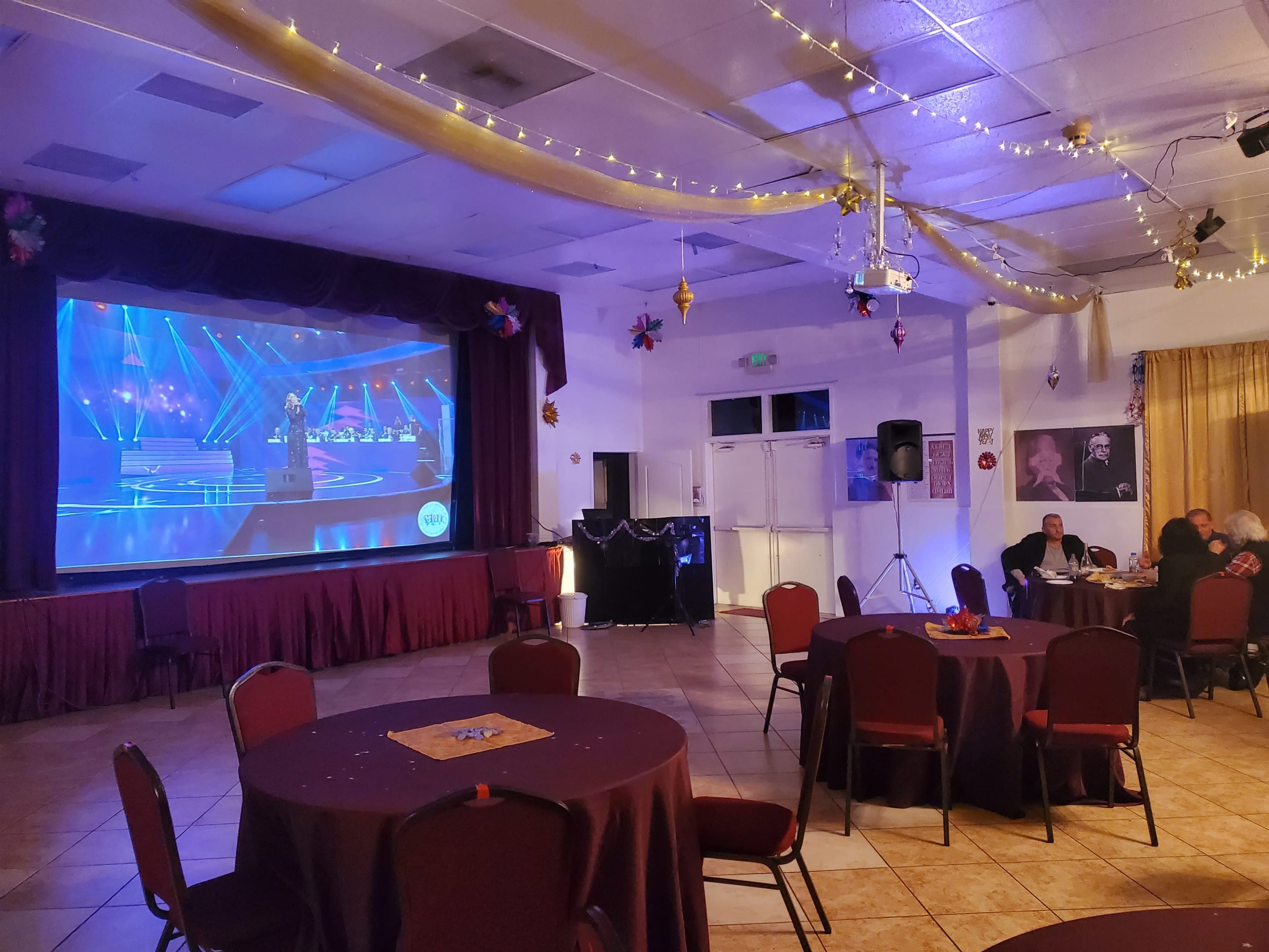 A performer stands on stage in front of a large screen displaying a musical performance, with round tables and chairs arranged in the foreground of the room.