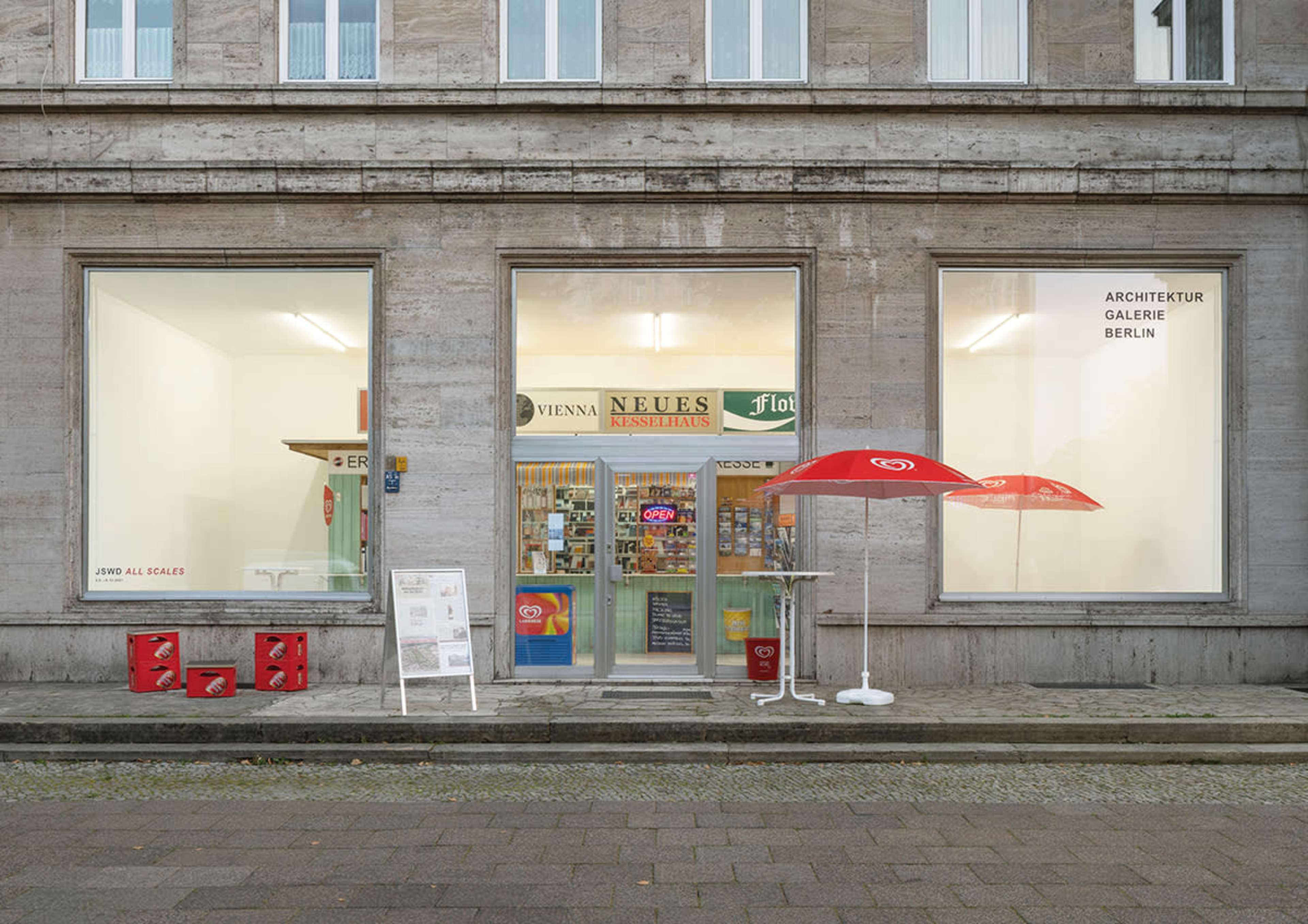 The exterior of a gallery storefront with large windows displaying an array of products and a red umbrella set outside.