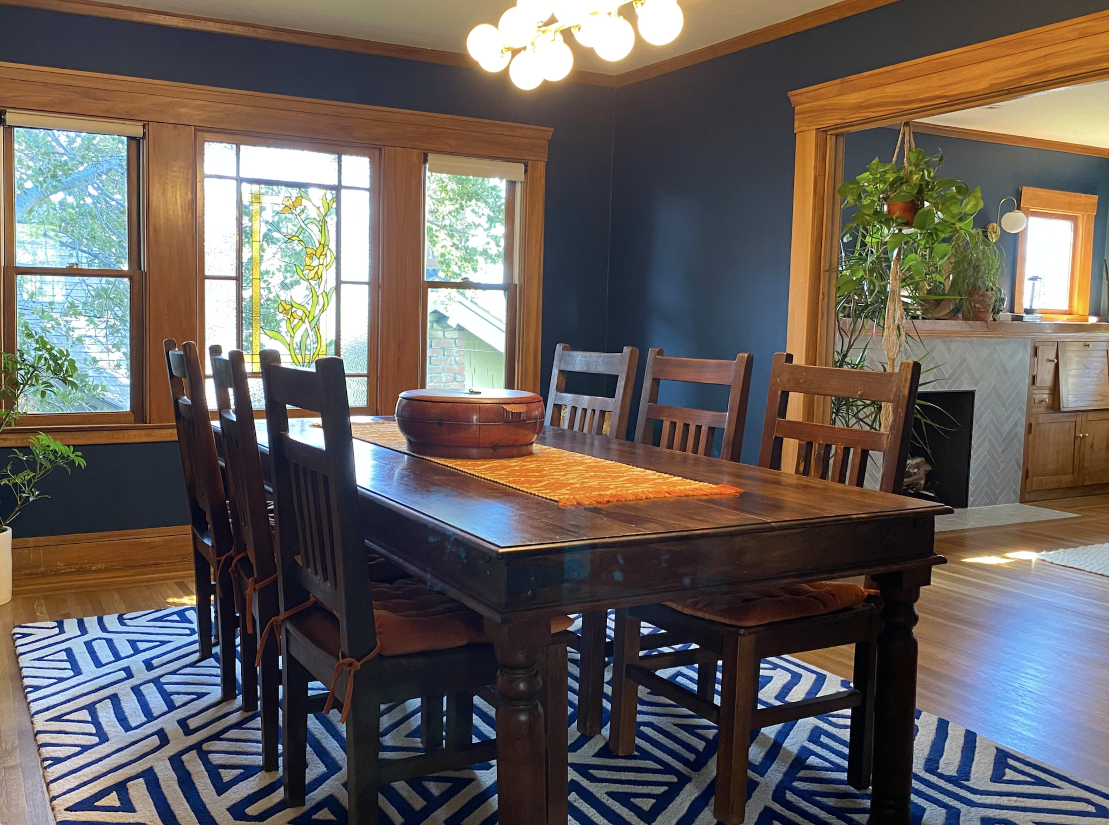 A wooden dining table with eight chairs is centered in a room featuring blue walls and large windows, with a patterned rug underneath.