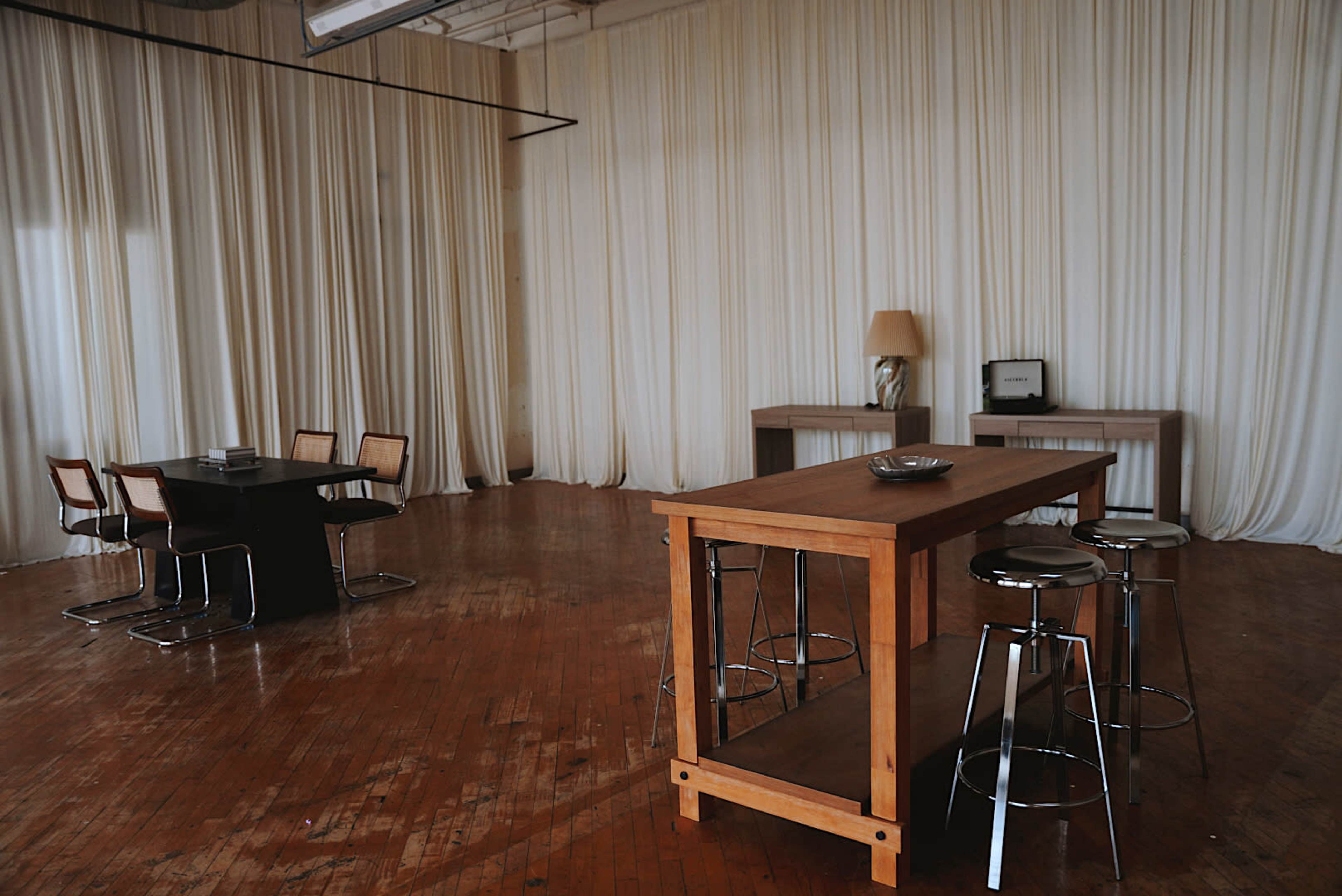 The photo shows a spacious interior with wooden flooring, a large table with stools, and a dining set in front of beige drapery.