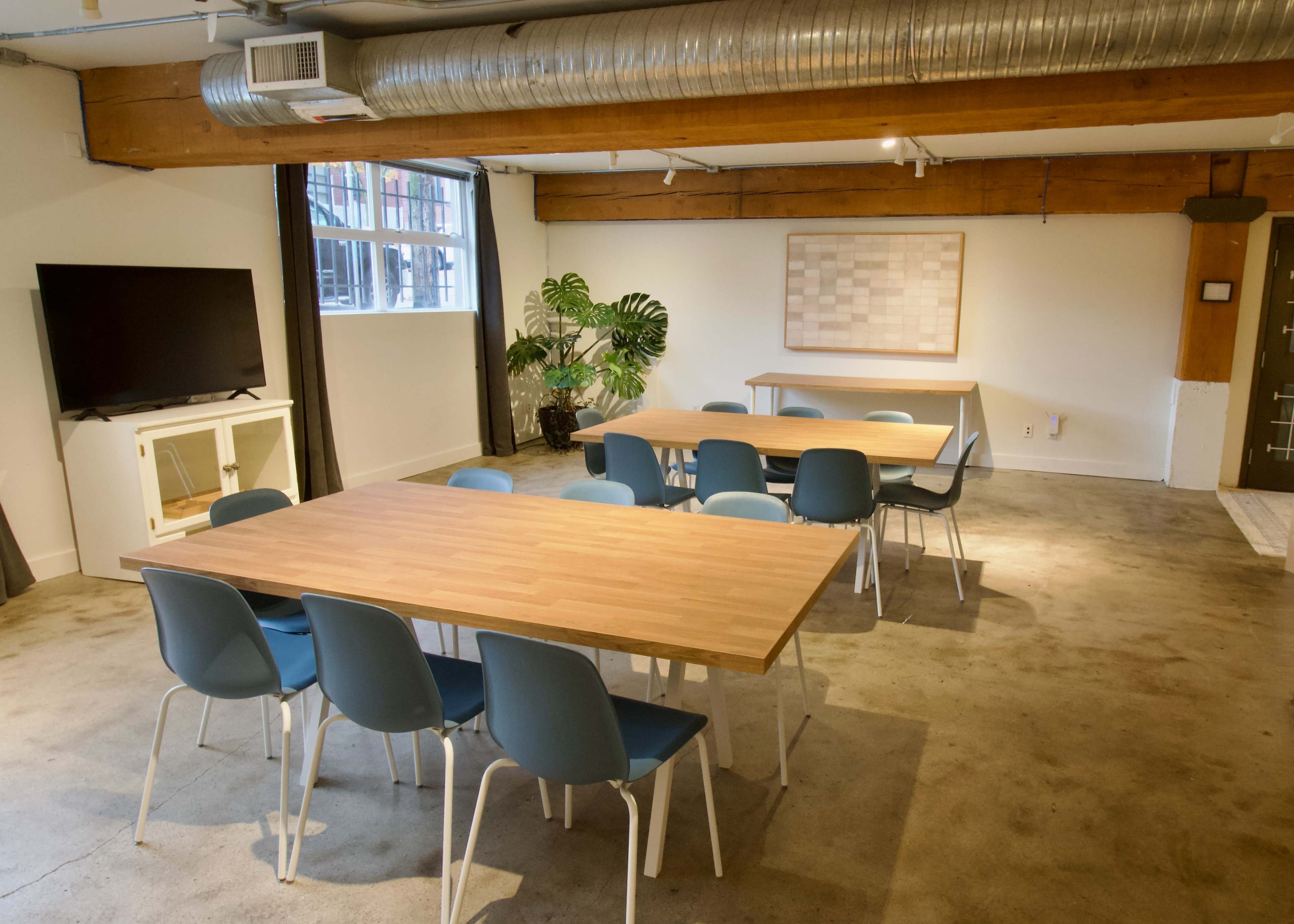 The image shows a modern conference room with two rectangular wooden tables surrounded by blue chairs, a large flat-screen TV on a stand, and a decorative plant in the corner.