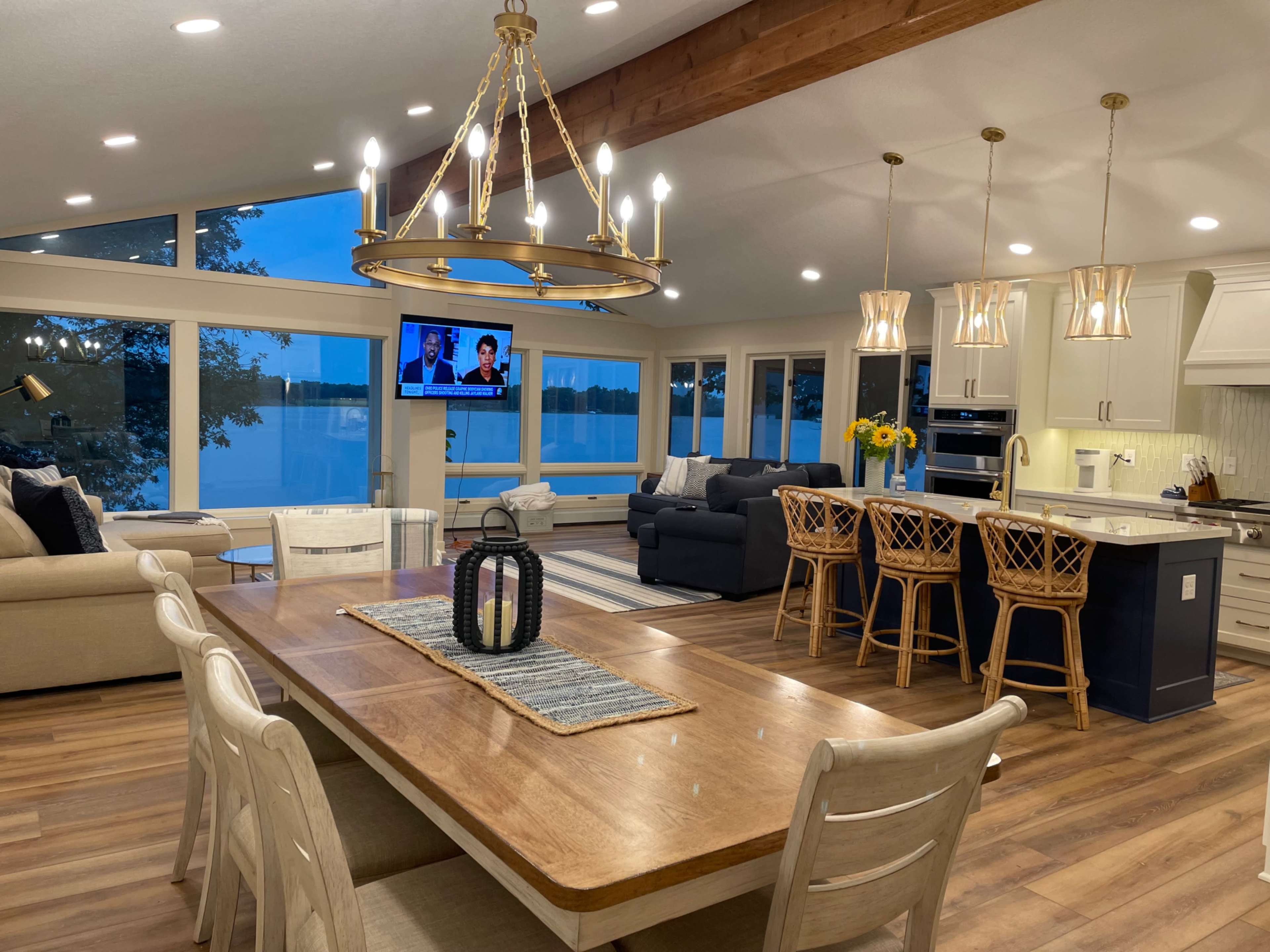 A modern living and dining area with large windows overlooking a body of water, featuring a dining table, comfortable seating, and a television mounted on the wall.