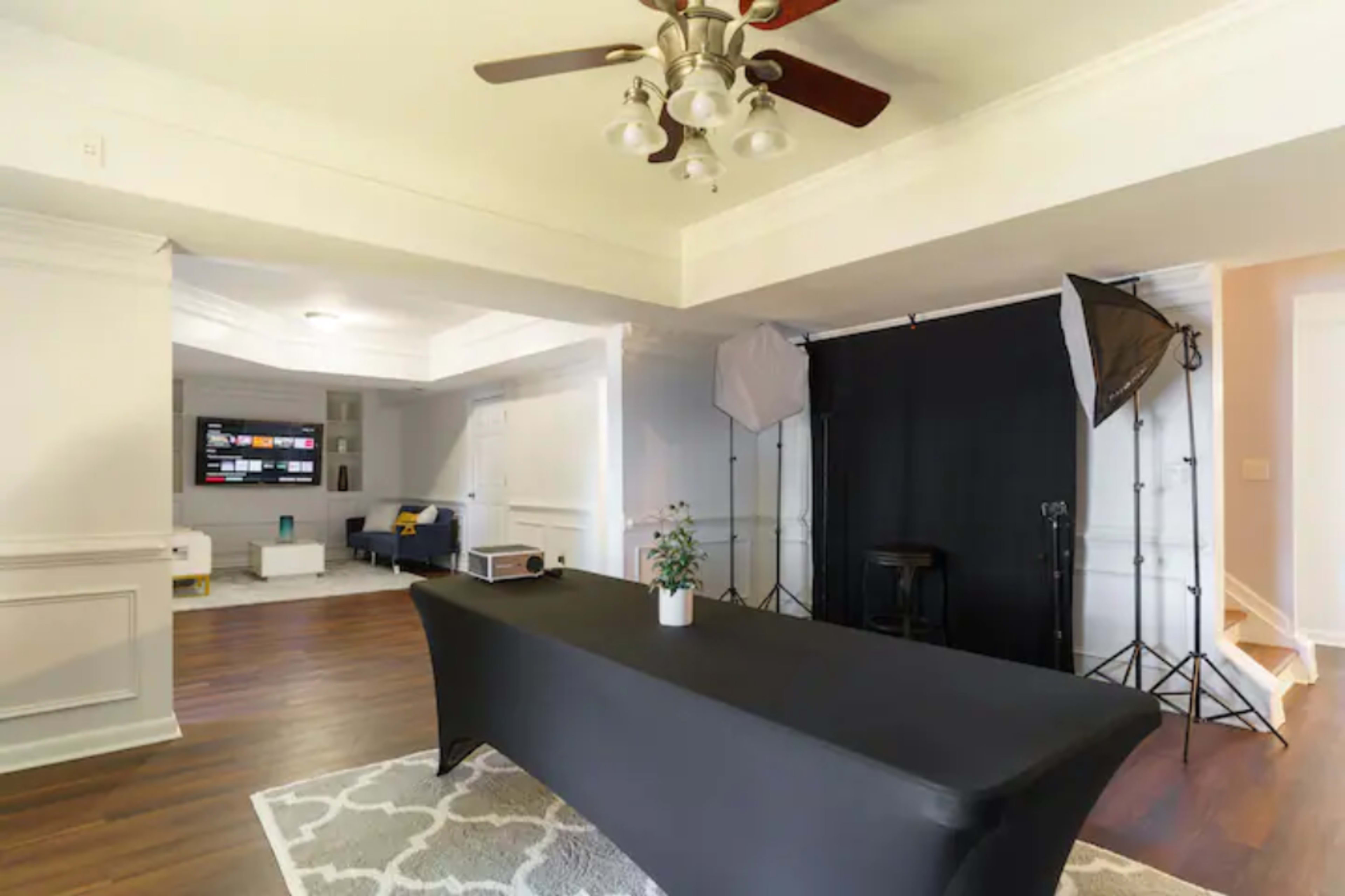 The image shows a spacious room with a dark table covered by a black tablecloth, studio lighting setup on one side, and a television mounted on a wall opposite a seating area.