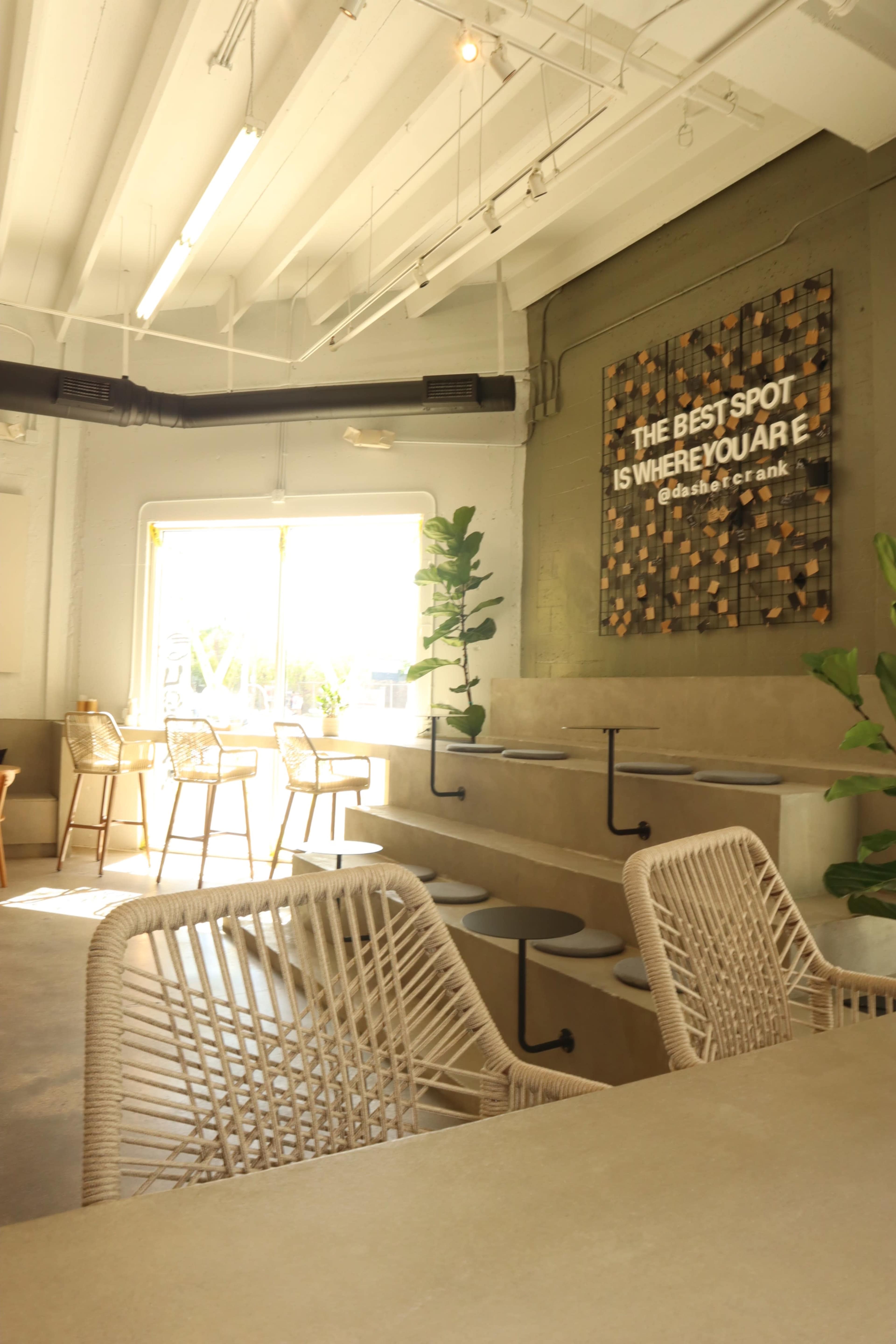 A modern café interior features a seating area with woven chairs, a concrete wall with a decorative quote, and large windows allowing natural light to fill the space.