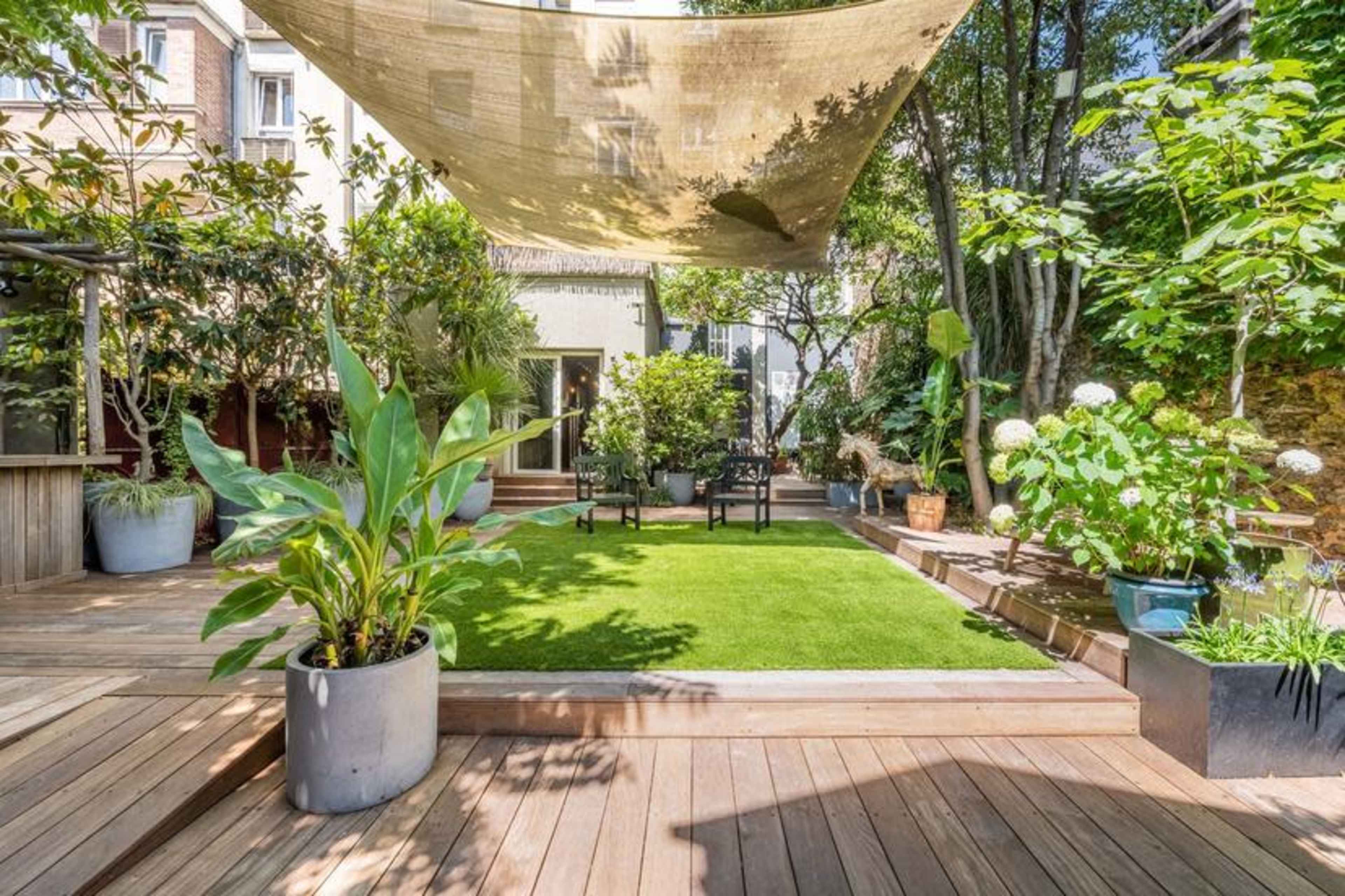 A landscaped garden area with wooden decking, featuring a green lawn, various plants in pots, and a shade cloth above seating arrangements.