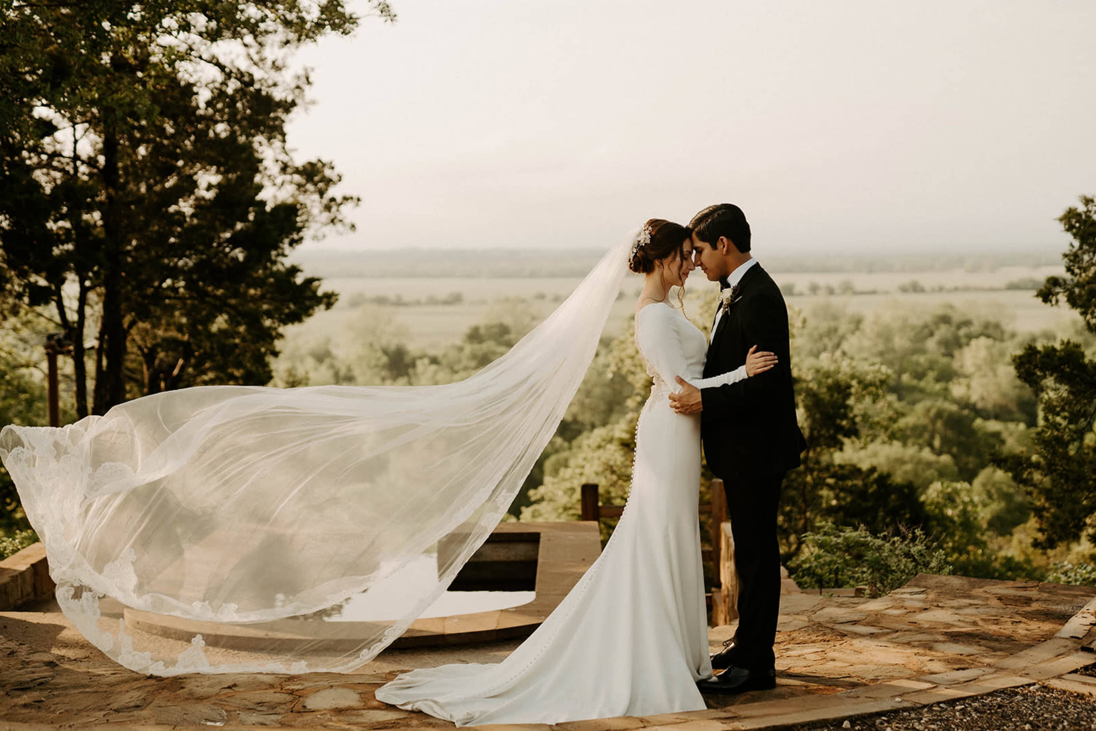 A couple stands embrace on a stone path with a flowing veil billowing in the breeze against a backdrop of lush greenery and a distant horizon.