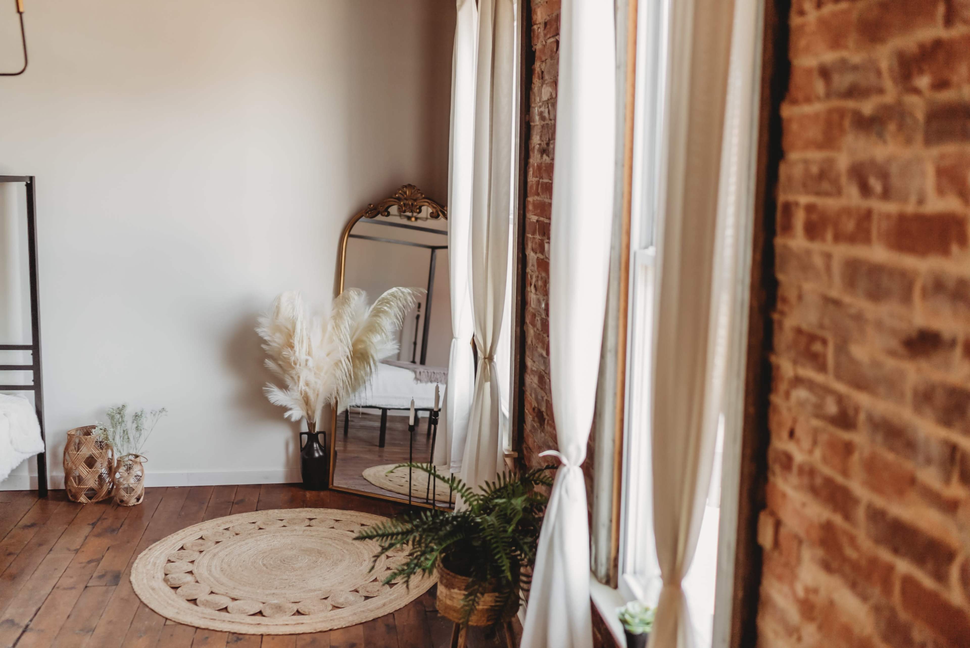 A well-lit room features a large mirror, a rattan rug, potted plants, and floor-to-ceiling windows with sheer curtains.
