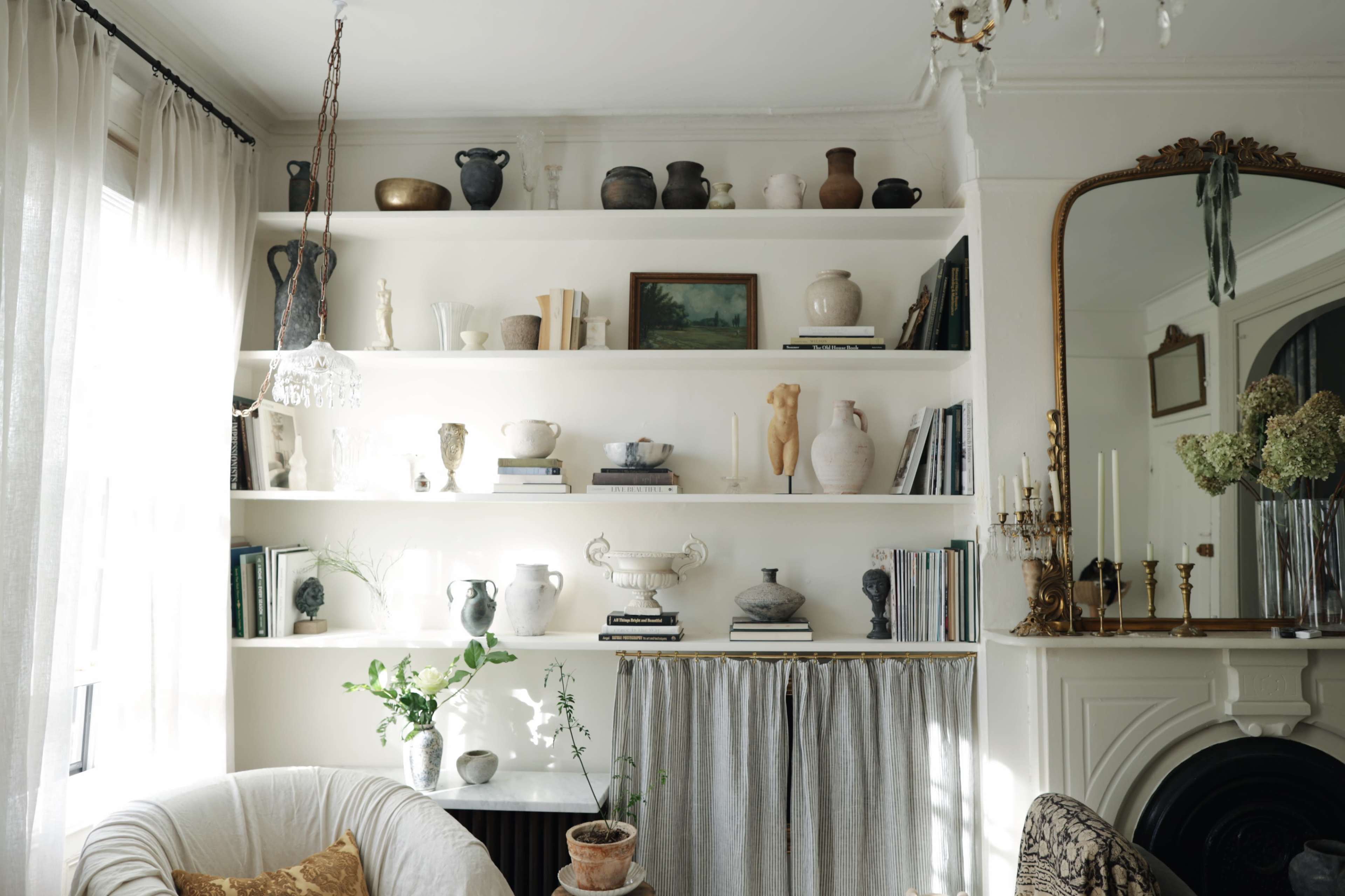 A well-organized shelf displays a variety of ceramic vases, books, and decorative items against a white wall in a softly lit room.