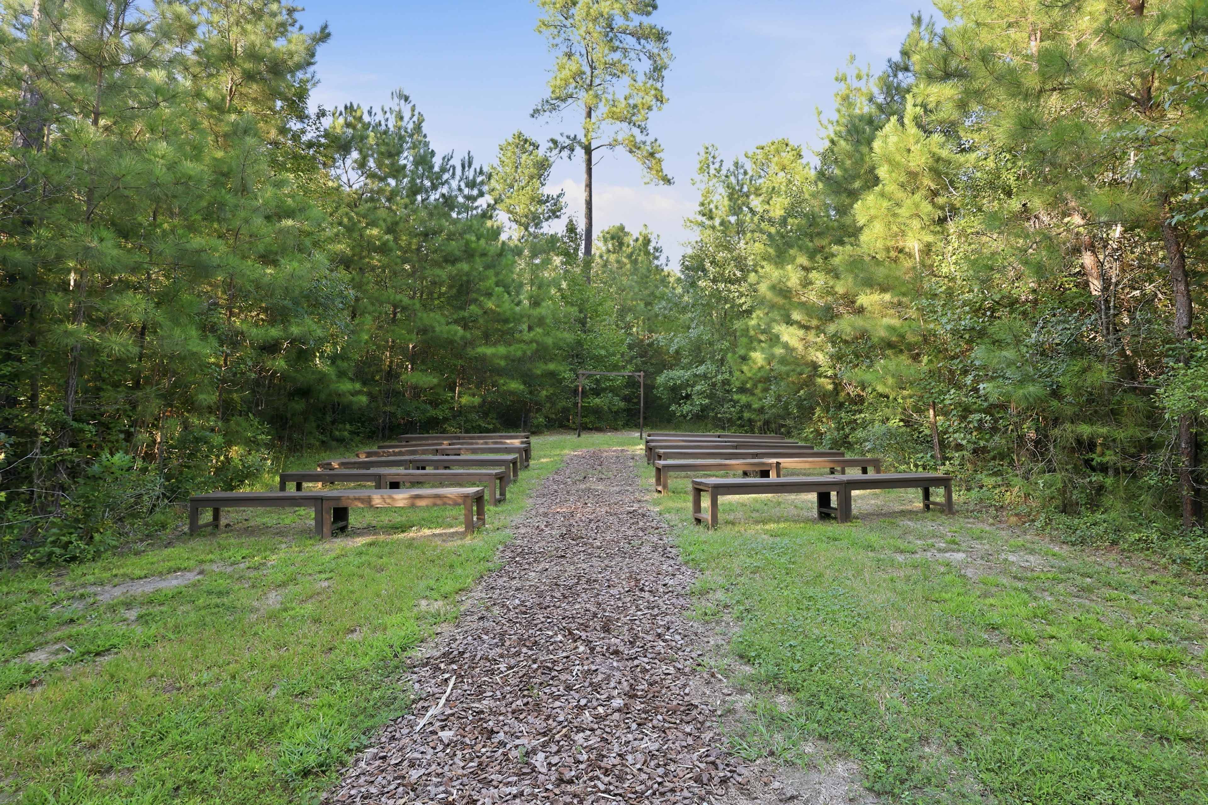 The image shows an outdoor meeting space with wooden benches arranged in rows along a path surrounded by trees and greenery.