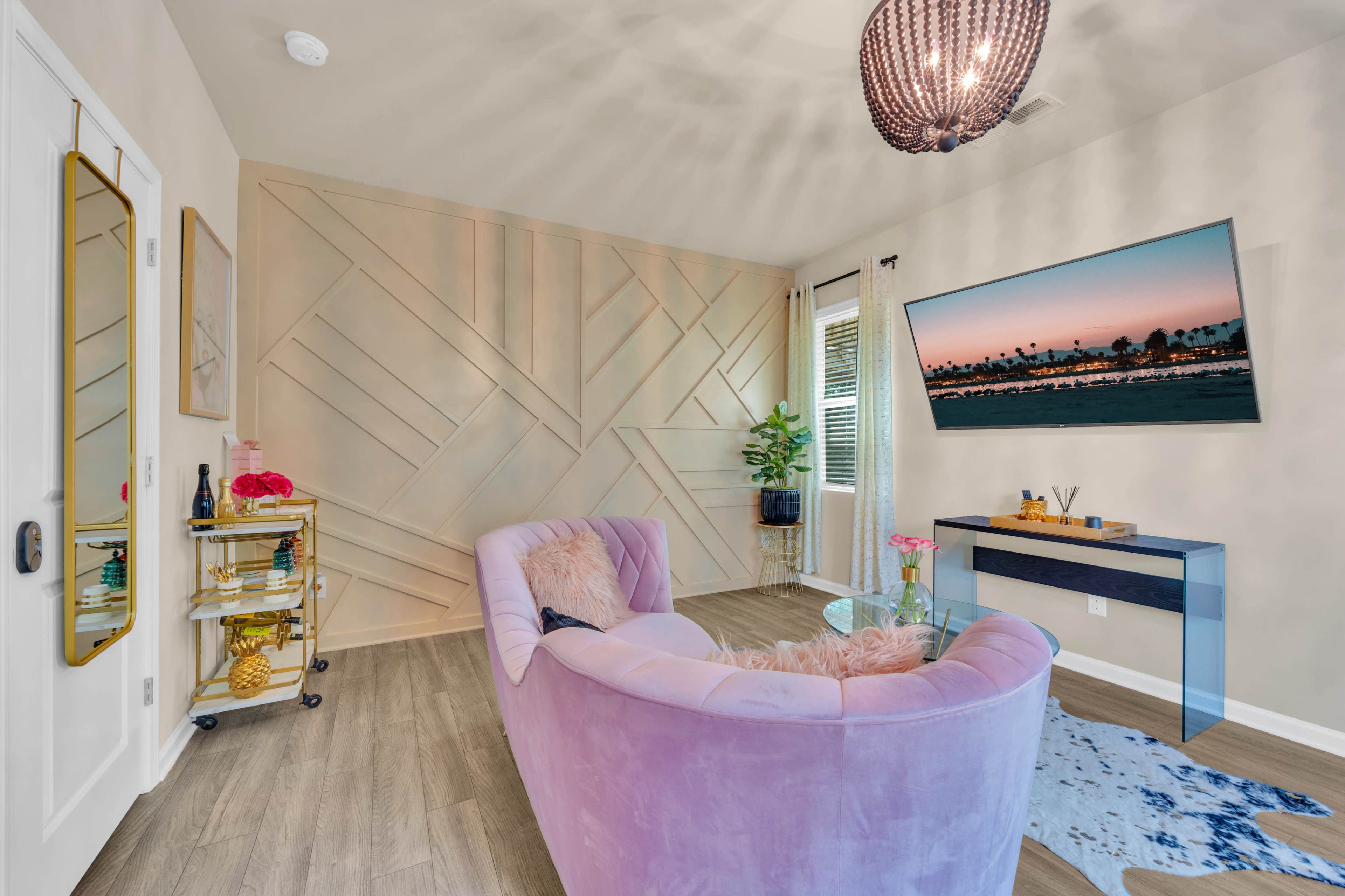A modern living room features a pink sofa, a geometric wall design, a television mounted on the wall, and a small bar cart next to a window.