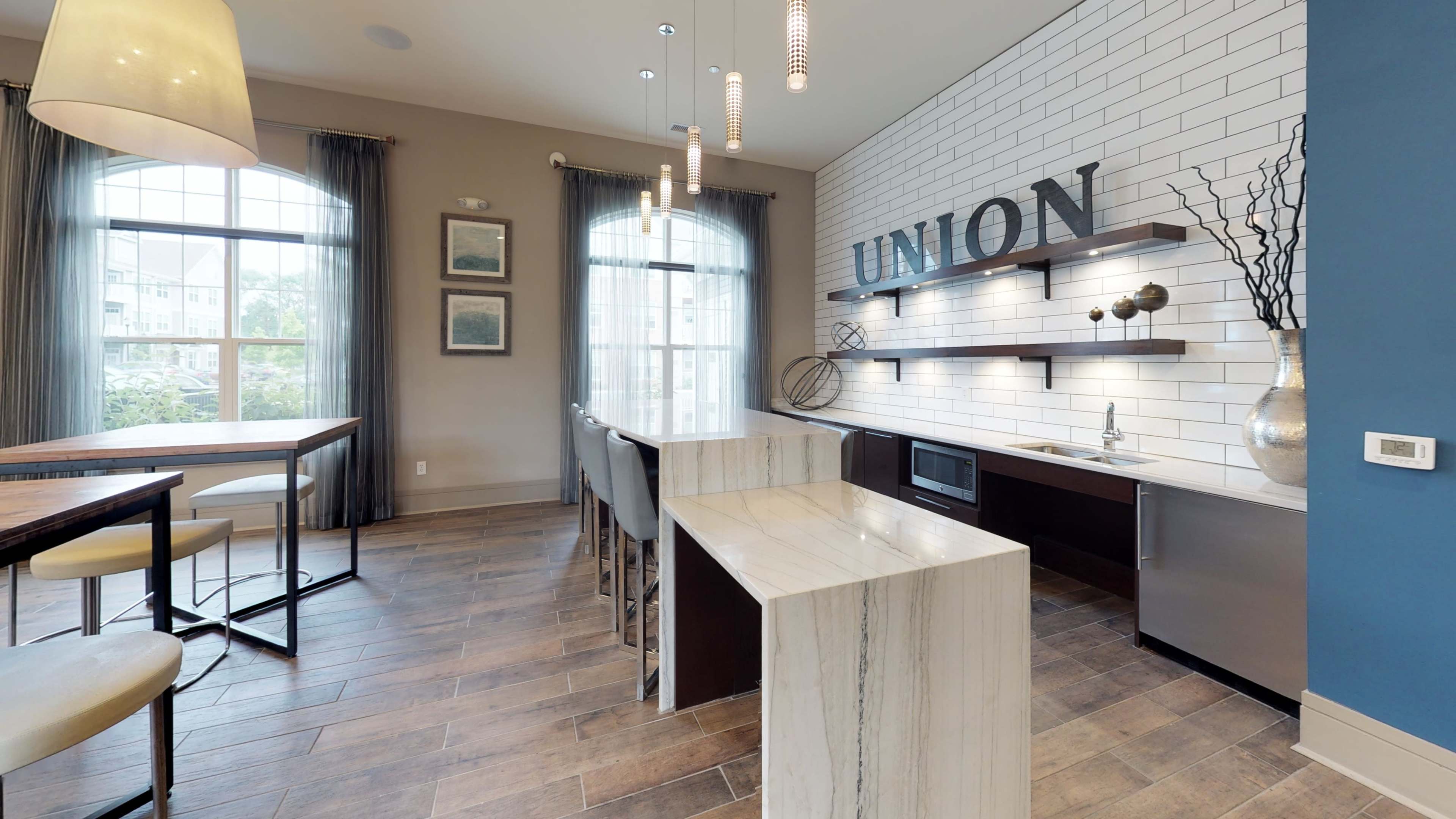 A modern kitchen area features a white tiled wall with "UNION" displayed, a countertop with a sink and microwave, and seating at a high table alongside large windows.