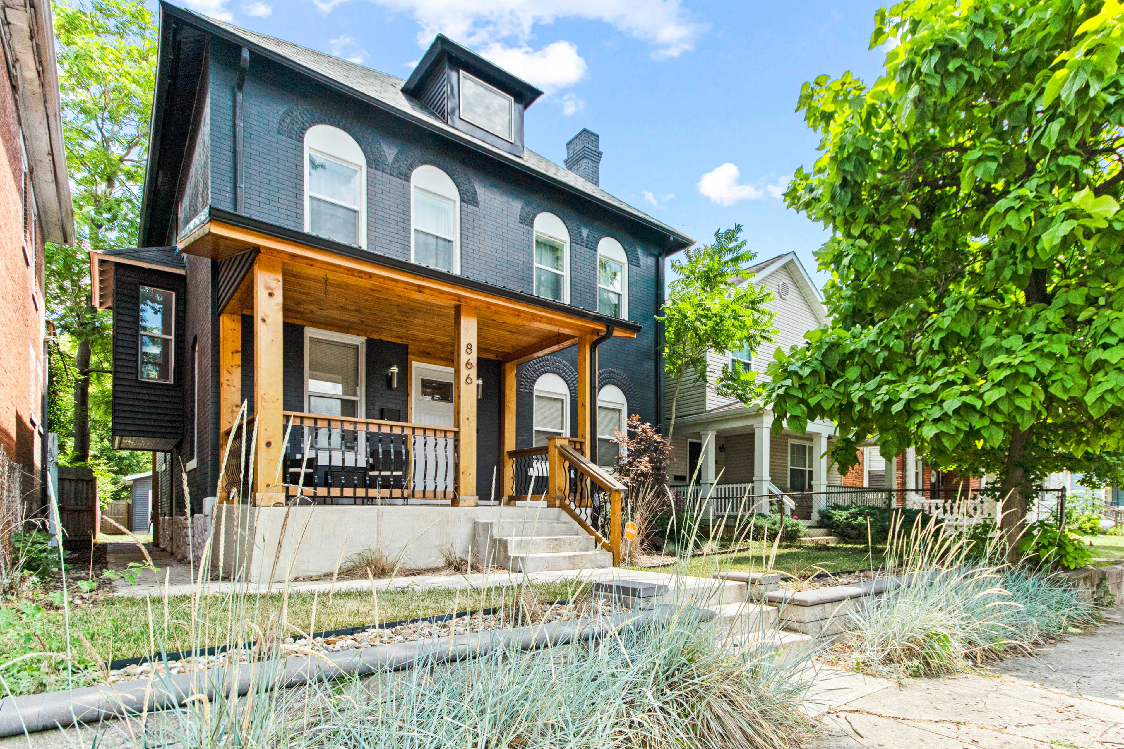 A dark-painted, three-story house with a wooden front porch and steps, surrounded by green foliage and other residential buildings.