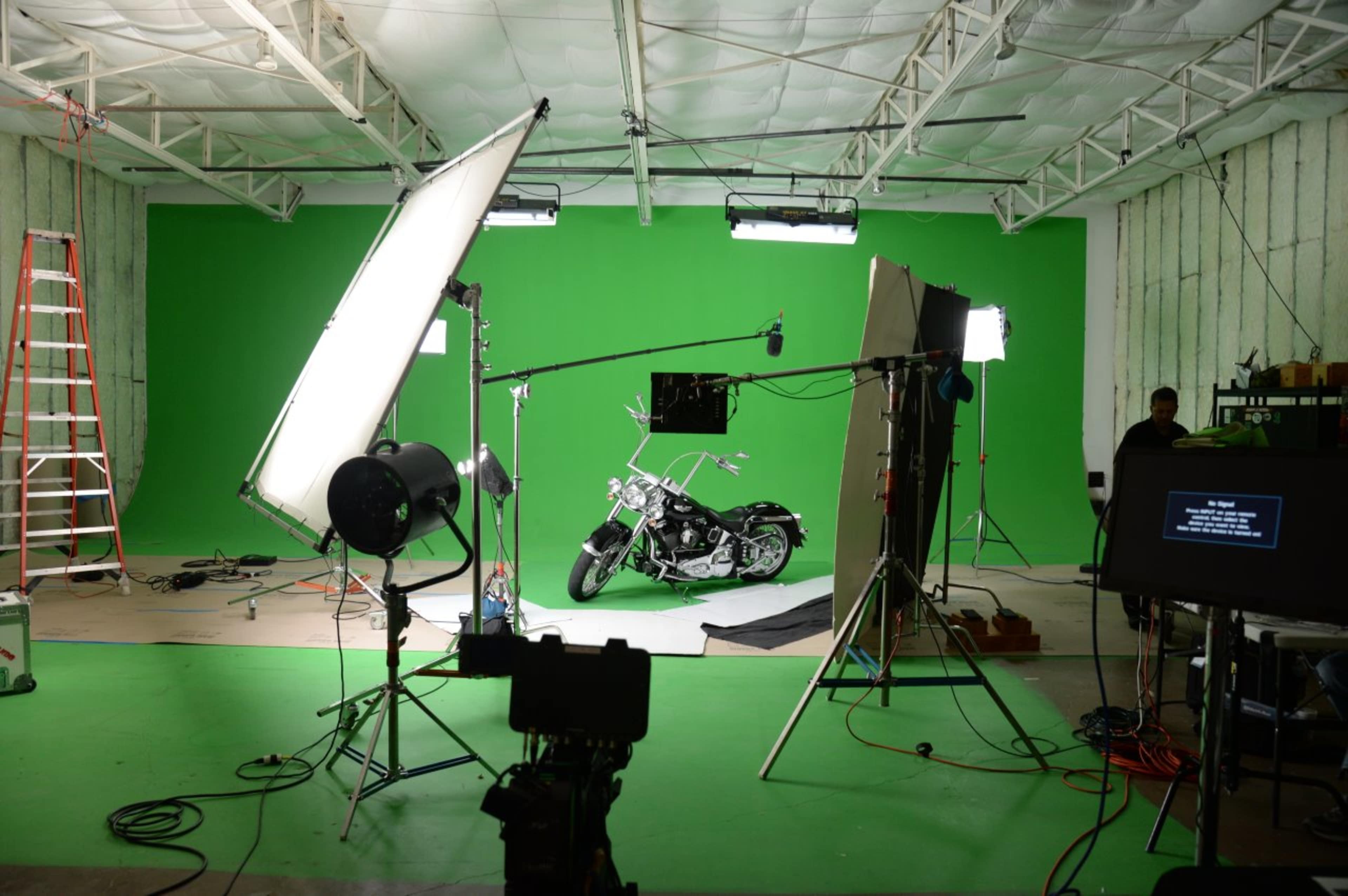 The image shows a motorcycle positioned on green flooring in a studio with green backdrop, surrounded by professional lighting and production equipment.