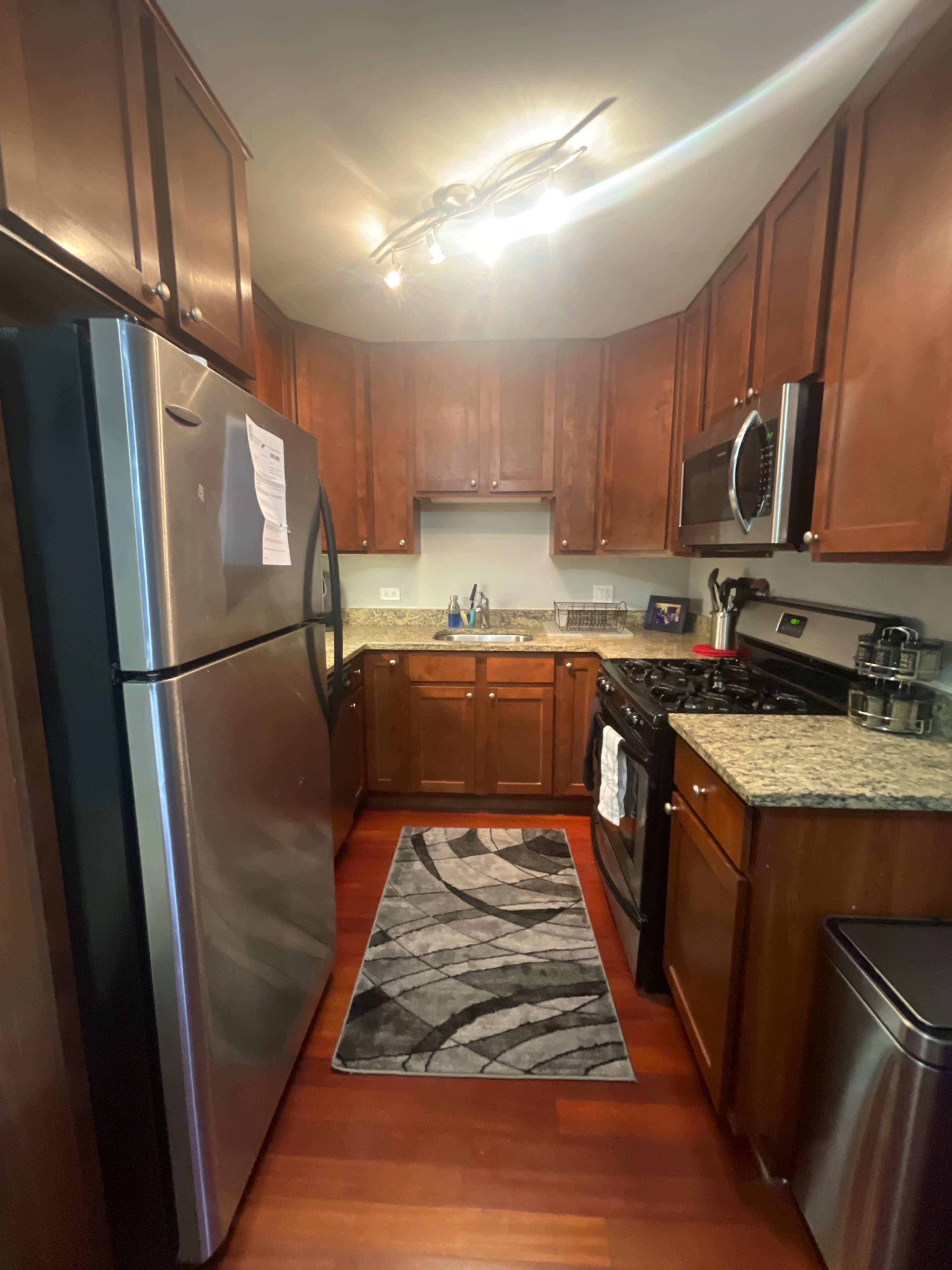 A kitchen features stainless steel appliances, wooden cabinets, granite countertops, and a patterned rug on the floor.