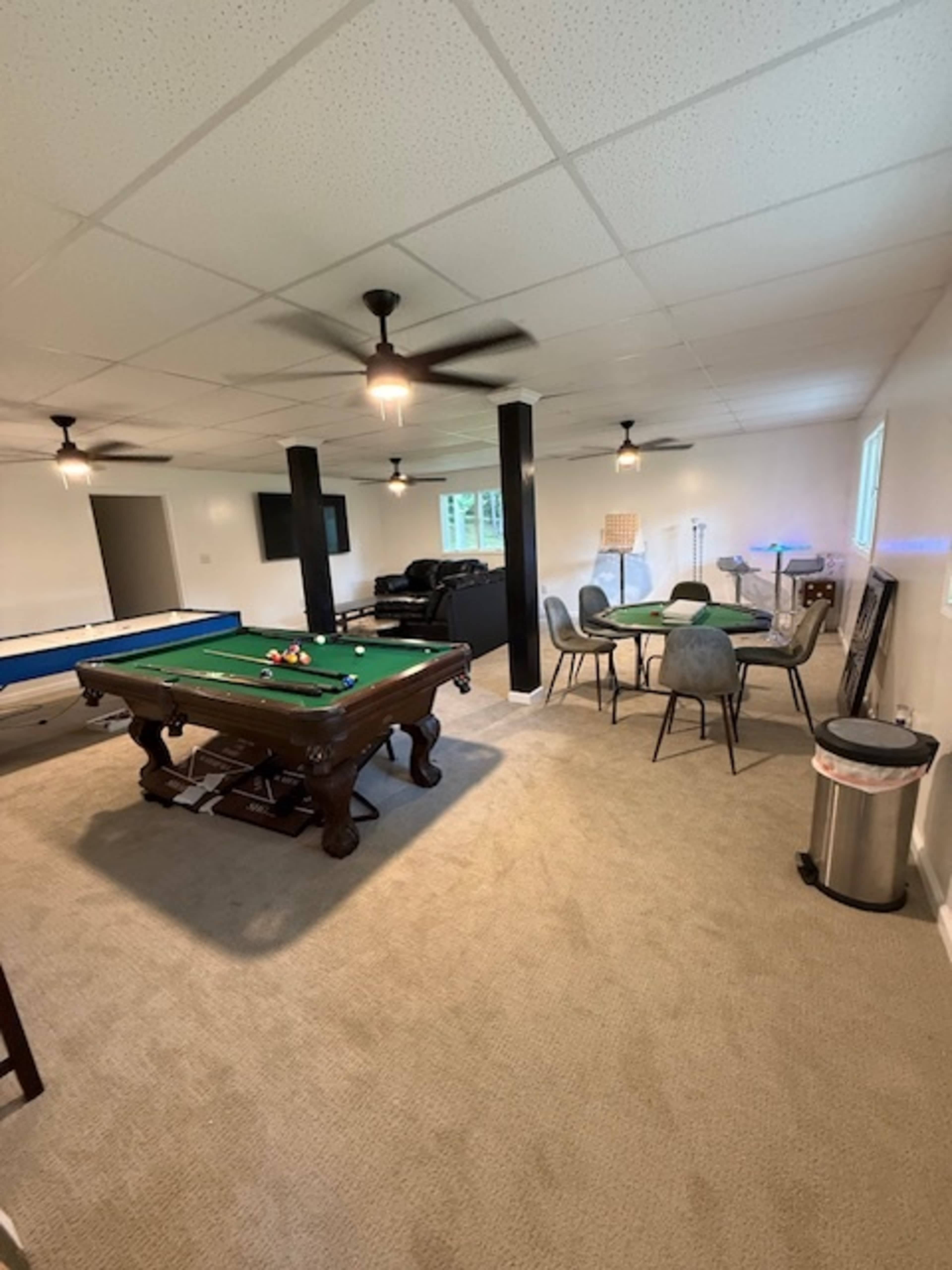 The image shows a well-lit basement recreation room featuring a pool table, a dining table with chairs, a sitting area, and a television.