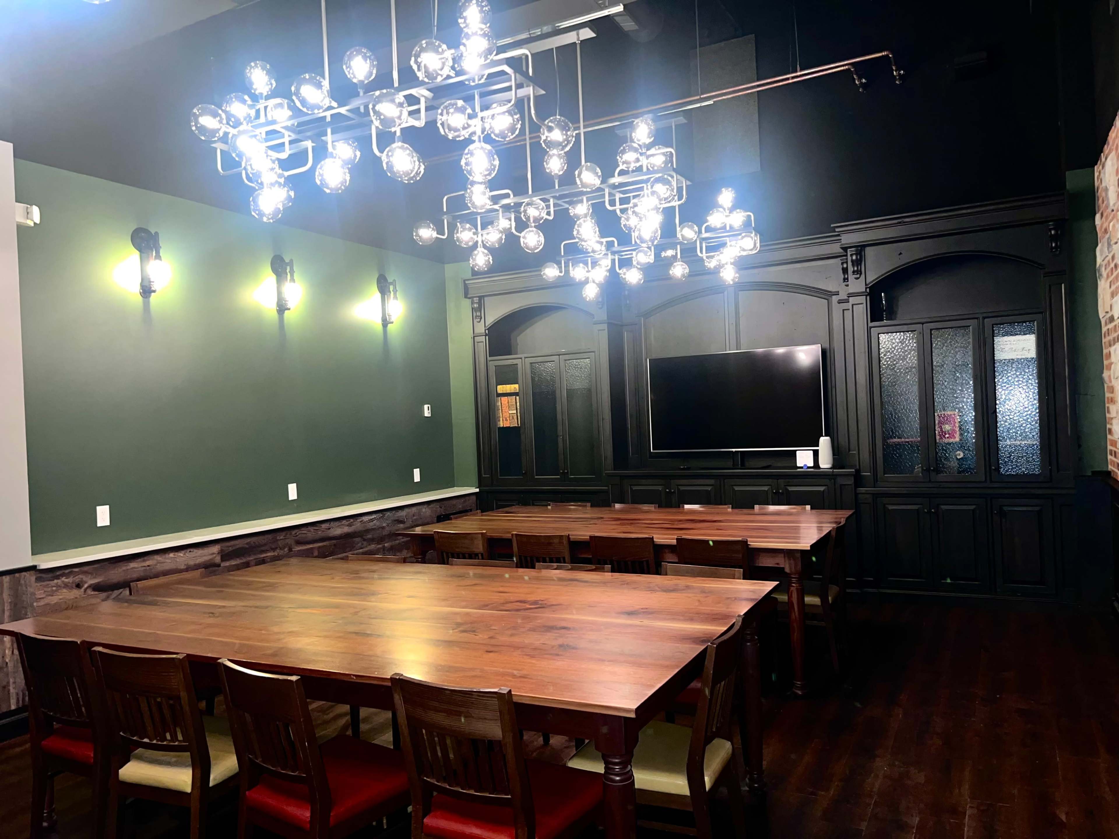 The image shows a conference room equipped with a long wooden table, chairs, a large television screen, and an elaborate chandelier providing overhead lighting.