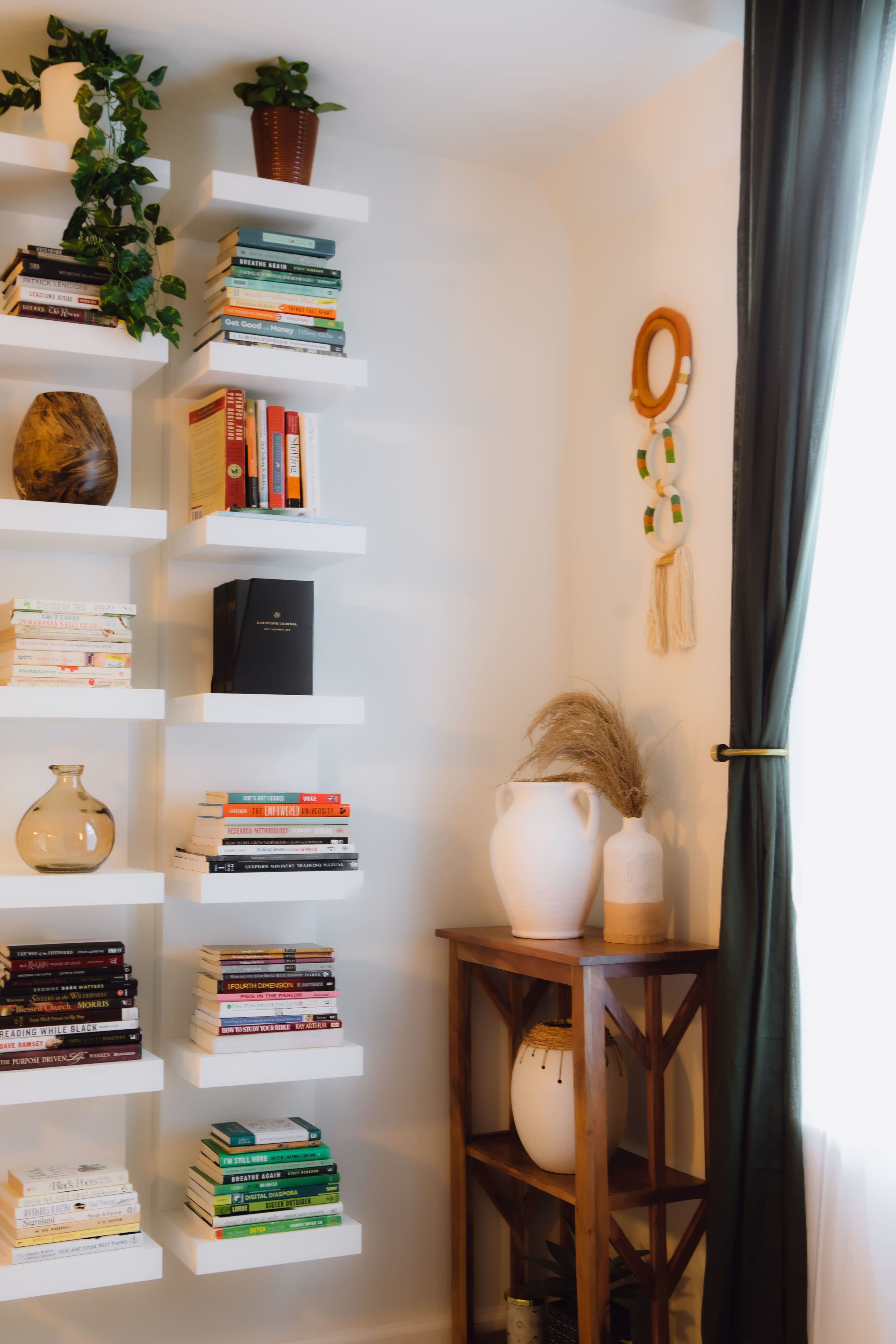 The image shows a corner of a room with white shelves filled with books, a potted plant, and decorative items including a vase and a woven wall hanging.