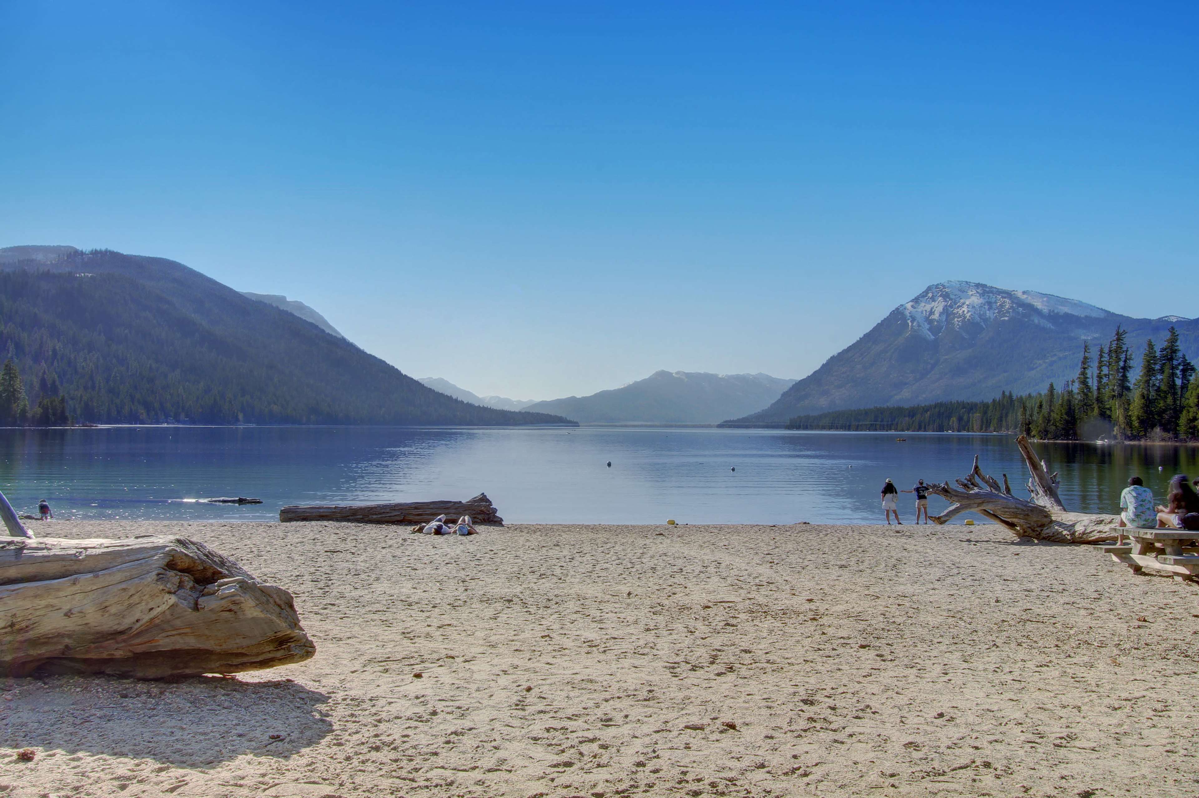 A sandy beach stretches along a calm lake surrounded by mountains and trees under a clear blue sky.