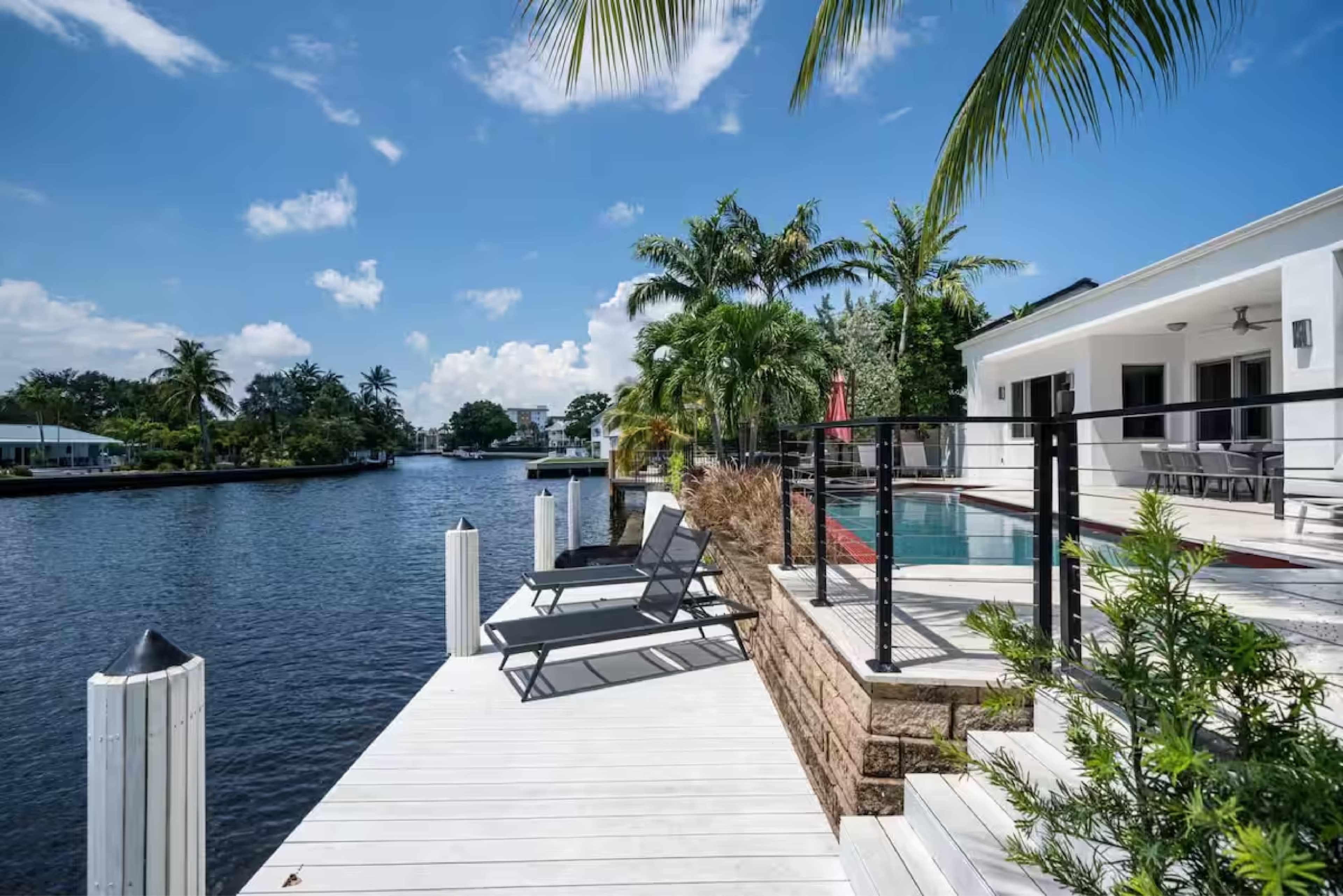 The image shows a waterfront property with a pool, lounge chairs, and tropical landscaping along a canal under a clear blue sky.