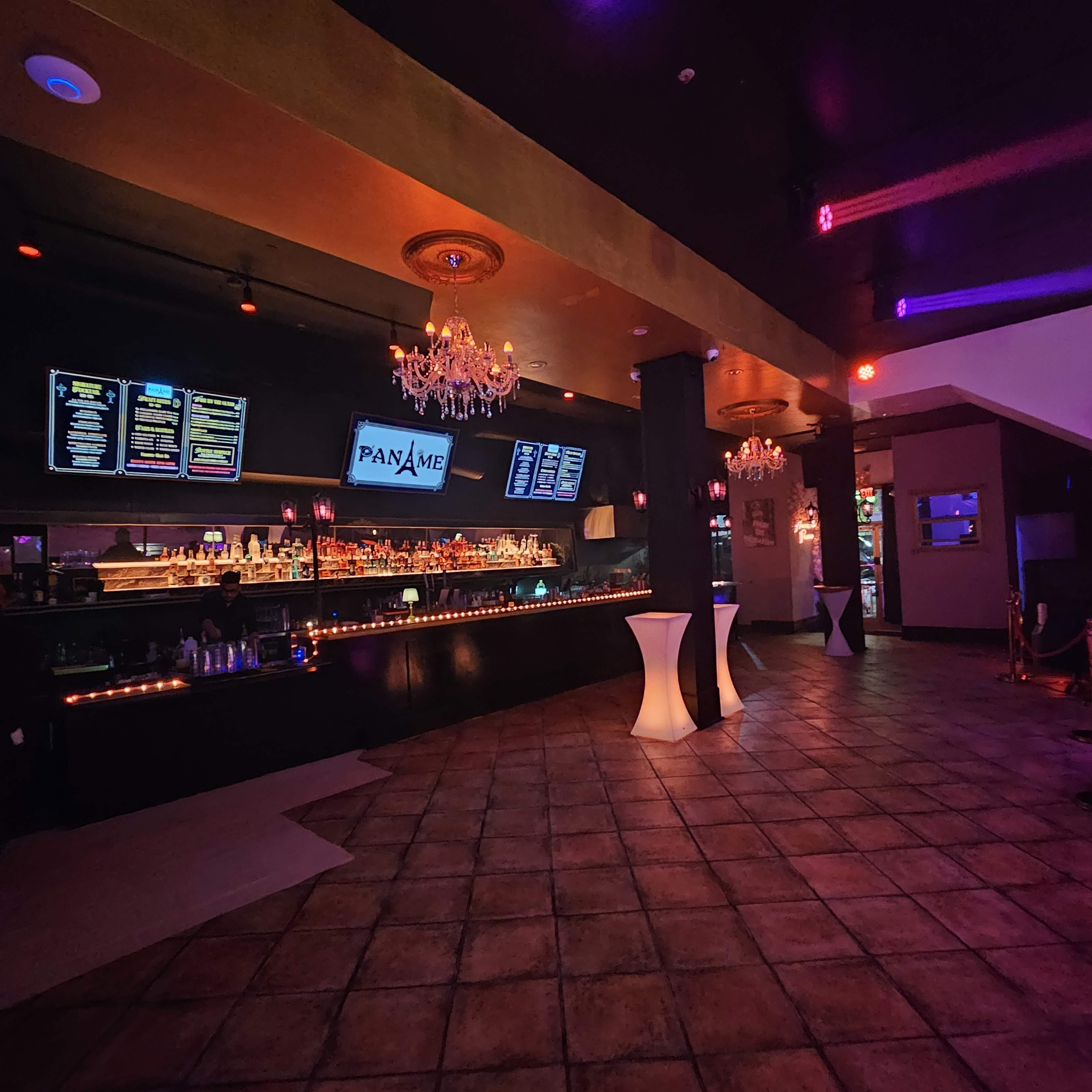 The image shows a dimly lit bar area with a long counter displaying various bottles, illuminated by colorful lights and decorative chandeliers.