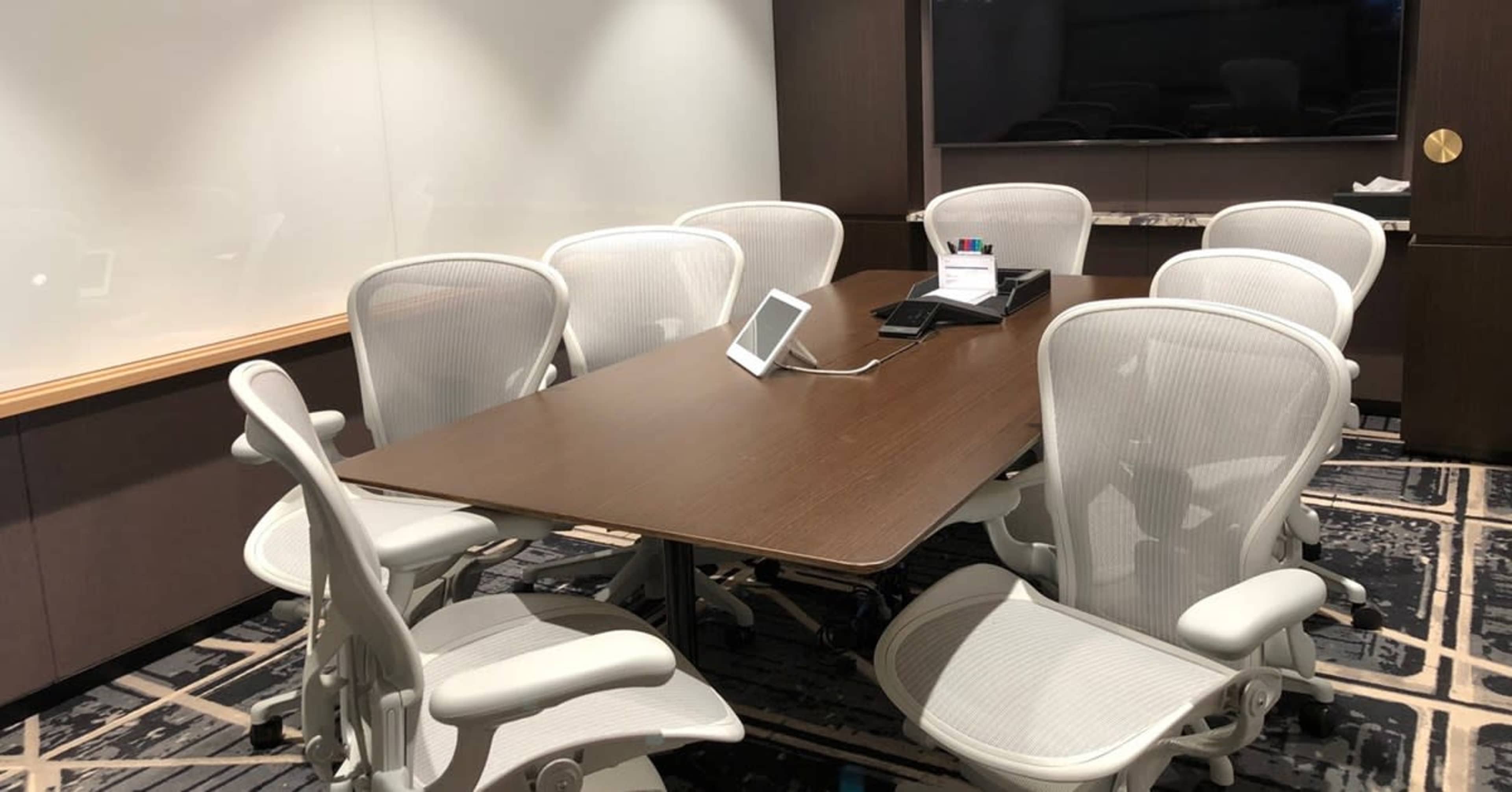 A conference room features a wooden table surrounded by several white office chairs and a screen on the wall.