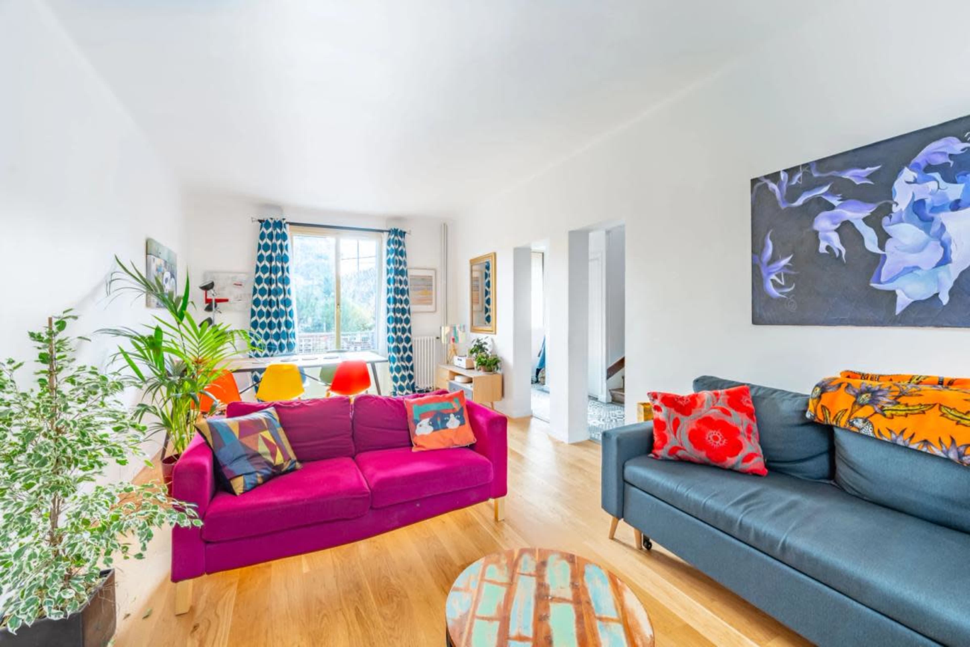 A bright living room features a pink sofa, a dark blue sofa, a round coffee table, and colorful artwork on the walls, with large windows letting in natural light.