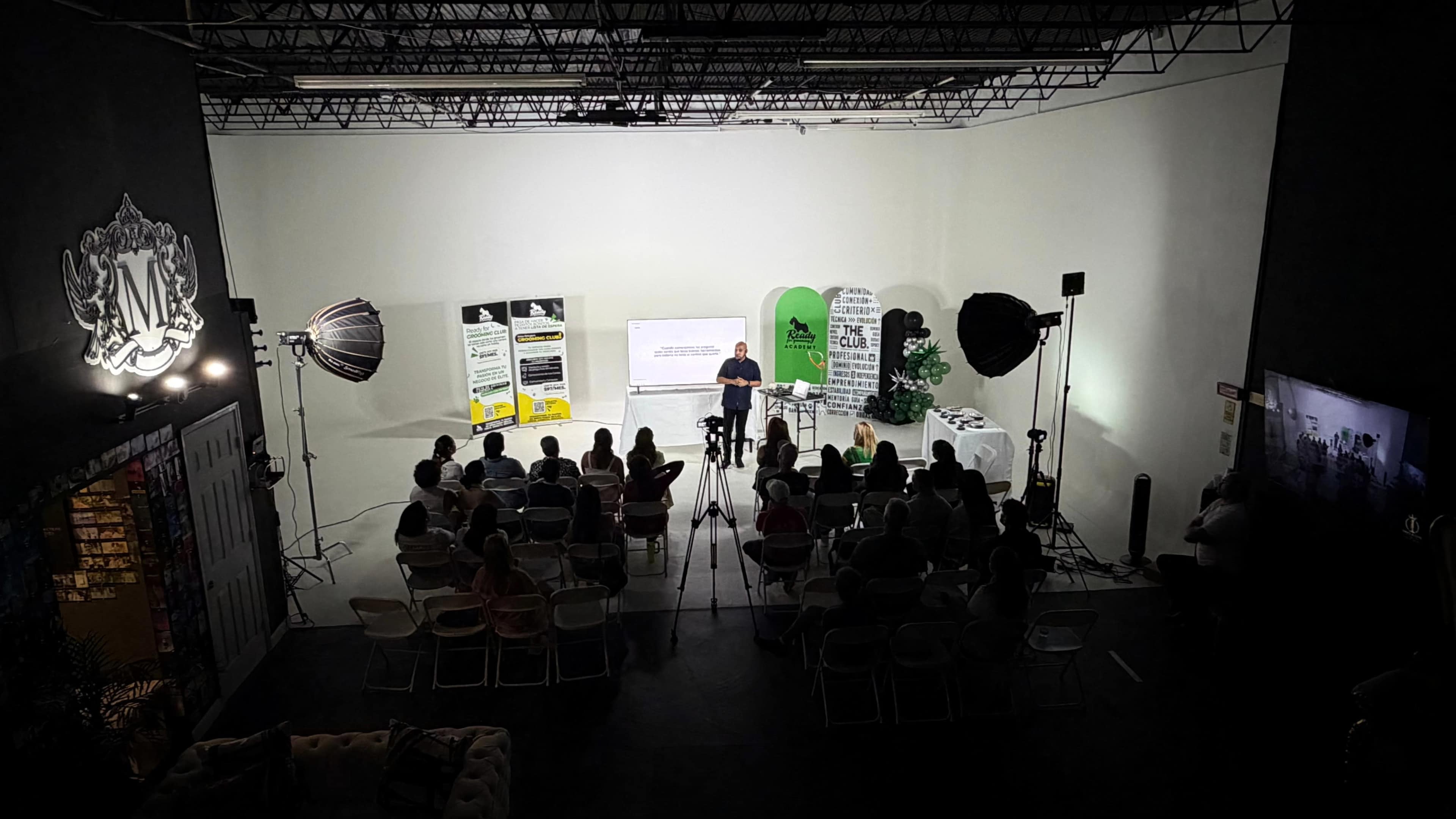 A speaker presents in front of an audience seated in a minimalist studio with various lighting equipment and informational displays.
